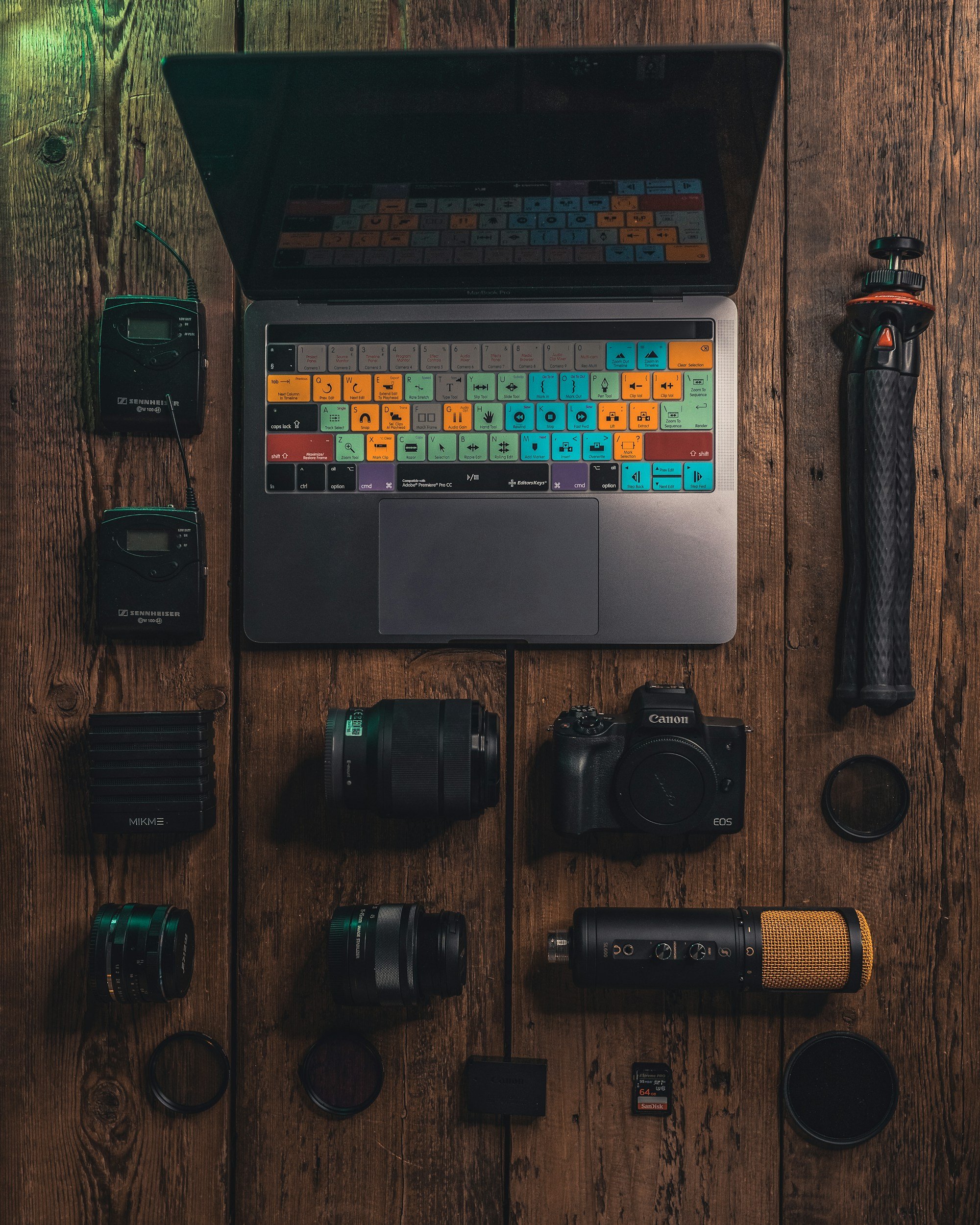 Photography equipment laid out on a wooden surface, including a laptop with colorful keyboard stickers, a camera, lenses, lenses filters, external microphones, SD card, memory card reader, and audio equipment.