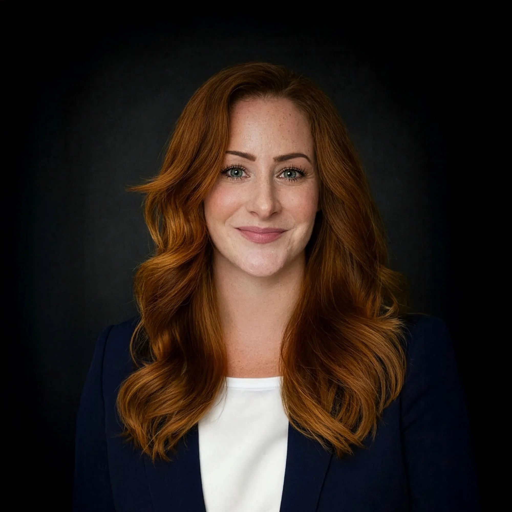 Headshot of a woman with long, wavy red hair, wearing a navy blazer and white top, smiling against a dark background.