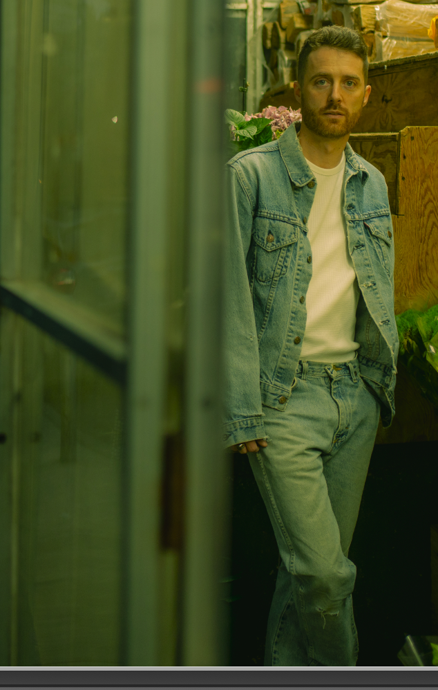 A man with short dark hair and a beard wearing a denim jacket and jeans, standing in a shop or warehouse with wooden shelves and plants in the background.