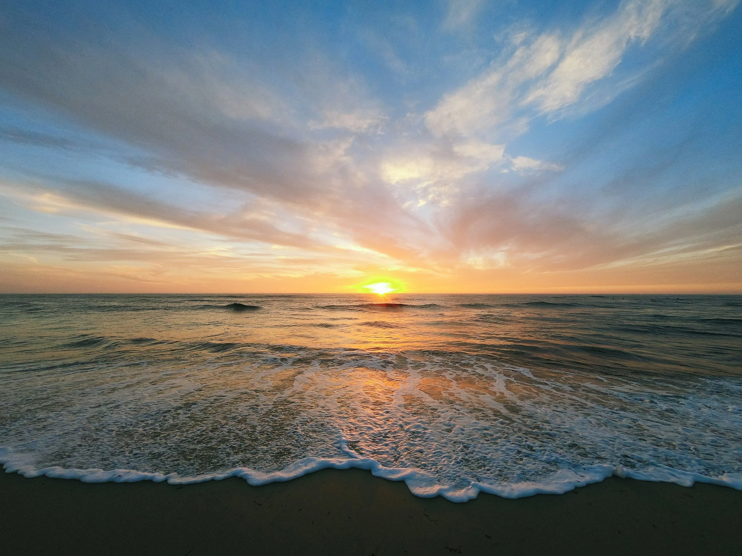 Sunset over the ocean with the sky filled with colorful clouds and gentle waves crashing on the sandy beach.