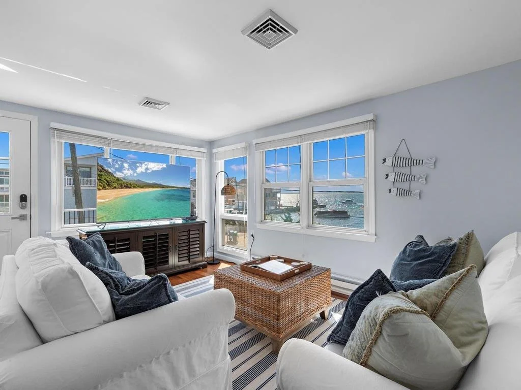 Living room with white sofas, throw pillows, a woven coffee table, a TV, and windows showing a seaside view.