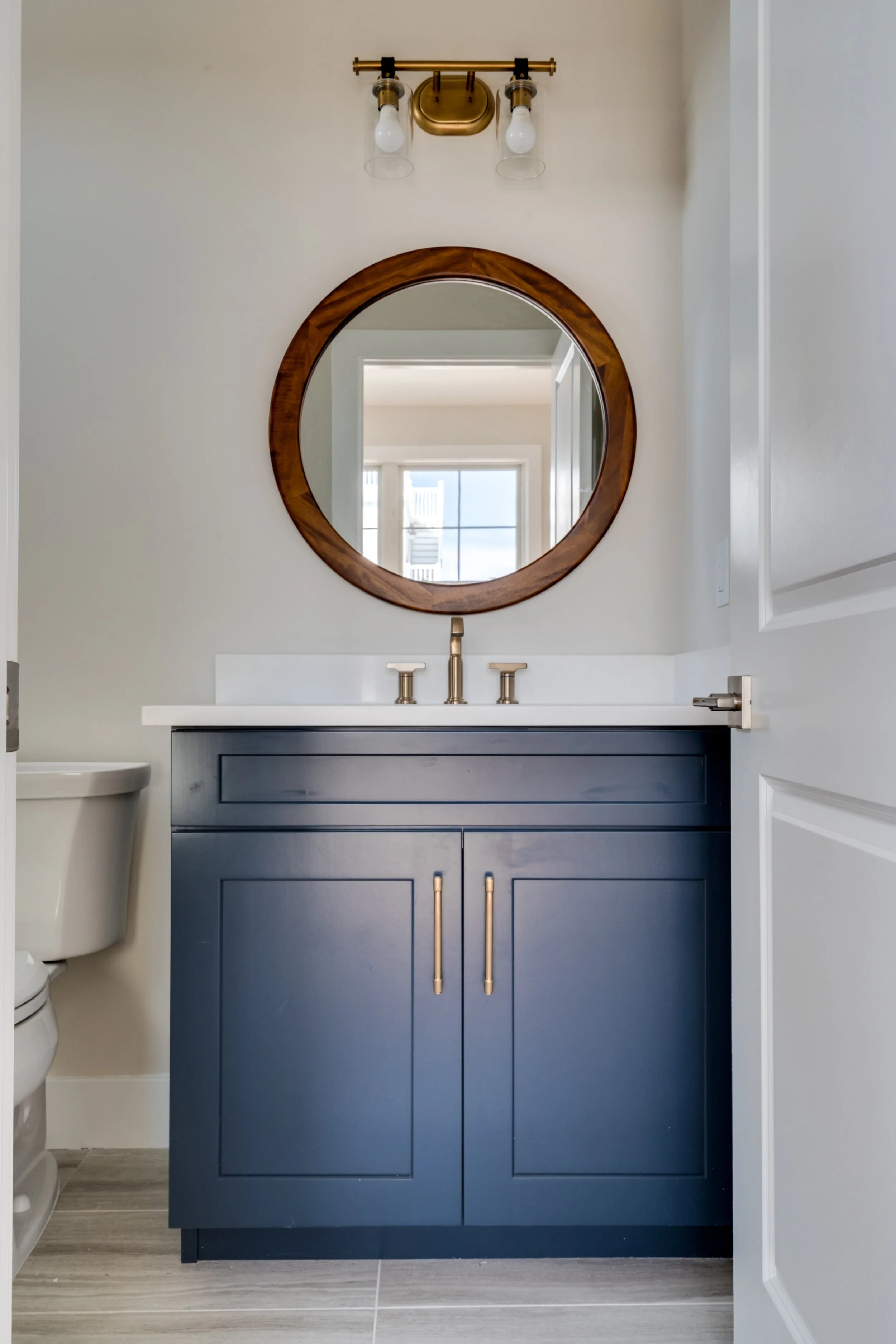 Bathroom vanity with navy blue cabinet, white countertop, round wooden mirror, and brass fixtures.