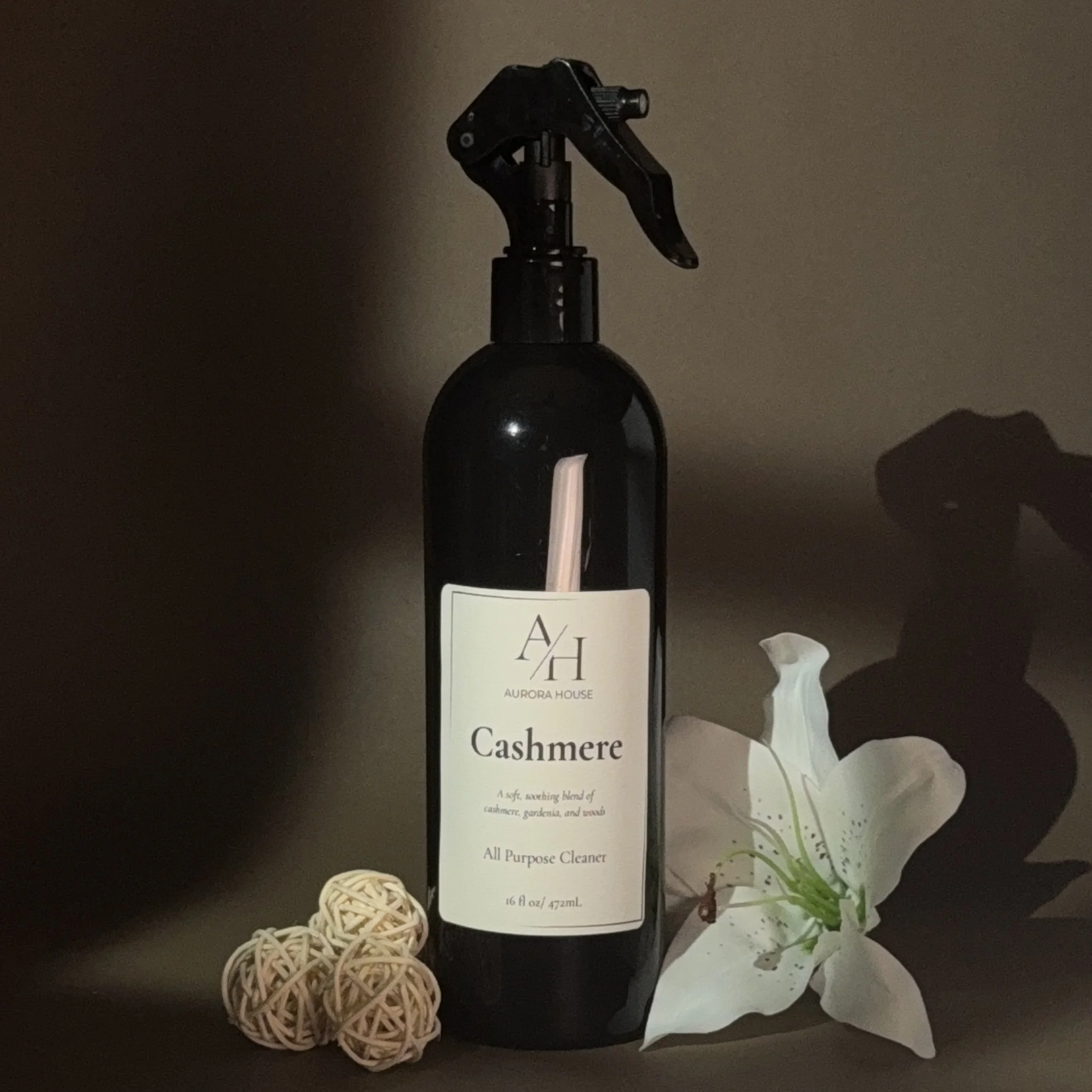 Black spray bottle labeled Aurora House Cashmere All Purpose Cleaner on a gray surface, with a white flower to the right and three decorative rattan balls to the left.