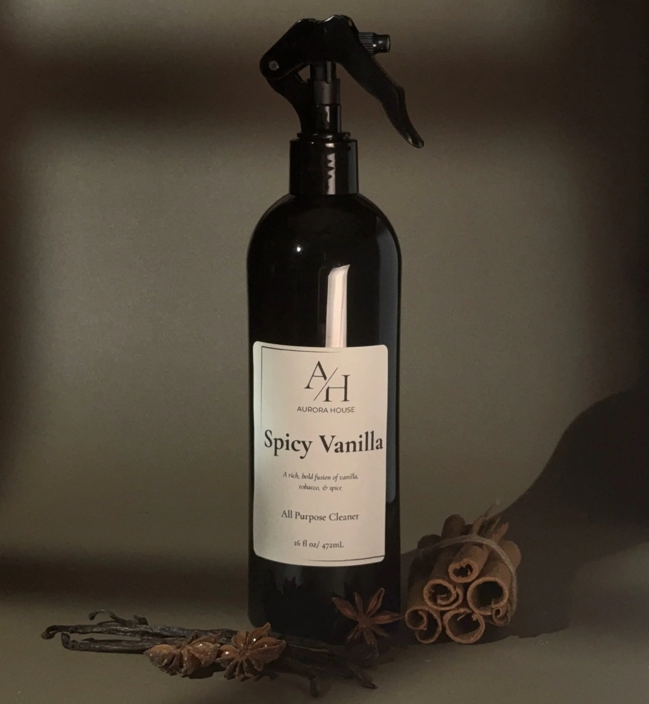 A black spray bottle labeled Aurora House Spicy Vanilla All Purpose Cleaner, with cinnamon sticks and star anise placed nearby on a gray surface against a beige background.