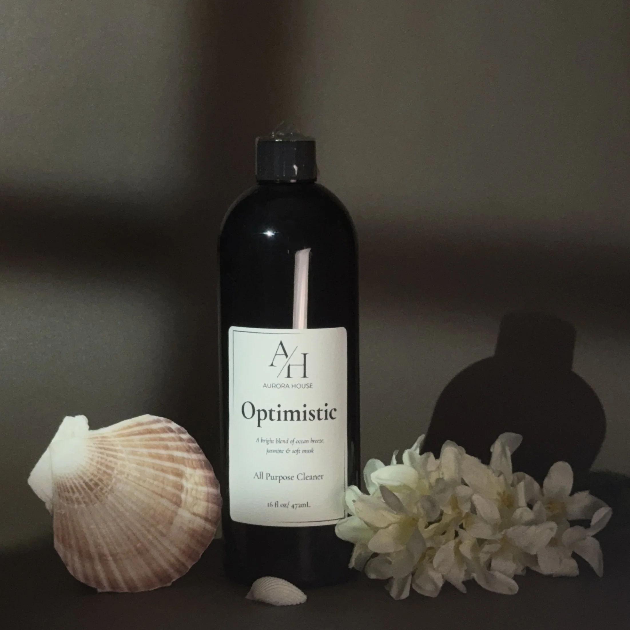 A black bottle labeled Aurora House Optimistic All Purpose Cleaner, 16 oz, with seashell and white flowers beside it, on a gray surface against a gray background.