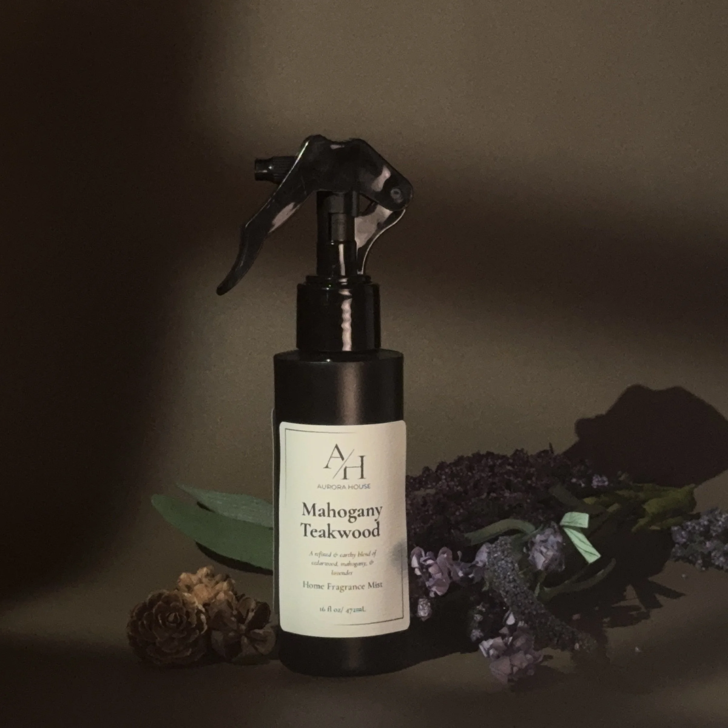 A black spray bottle labeled Aurora House Mahogany Teakwood Home Fragrance Mist on a wooden surface, with pinecones and purple lavender flowers beside it, against a textured beige background.