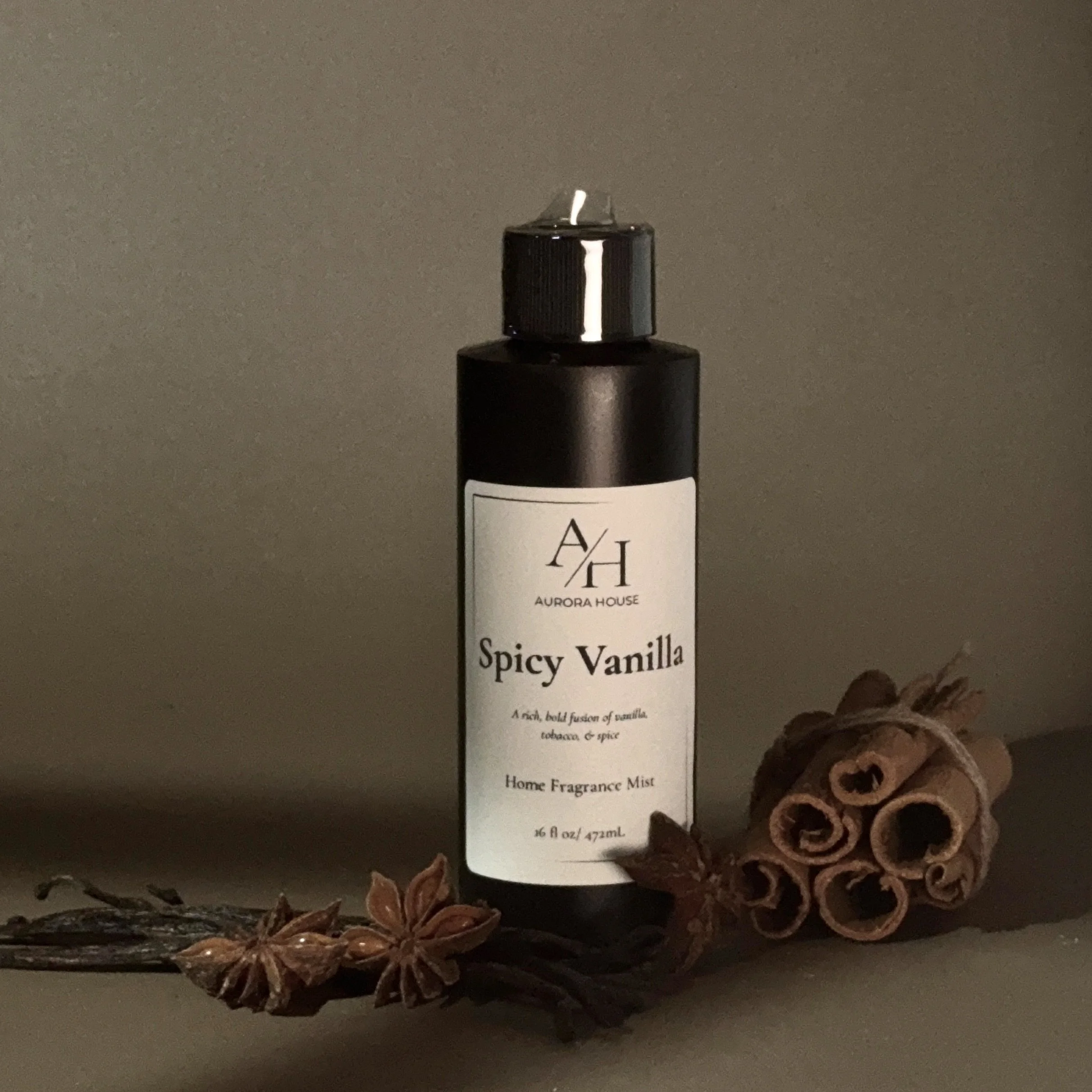 A black bottle of Aurora House Spicy Vanilla Home Fragrance Mist with cinnamon sticks and star anise spices placed in front of it on a neutral background.