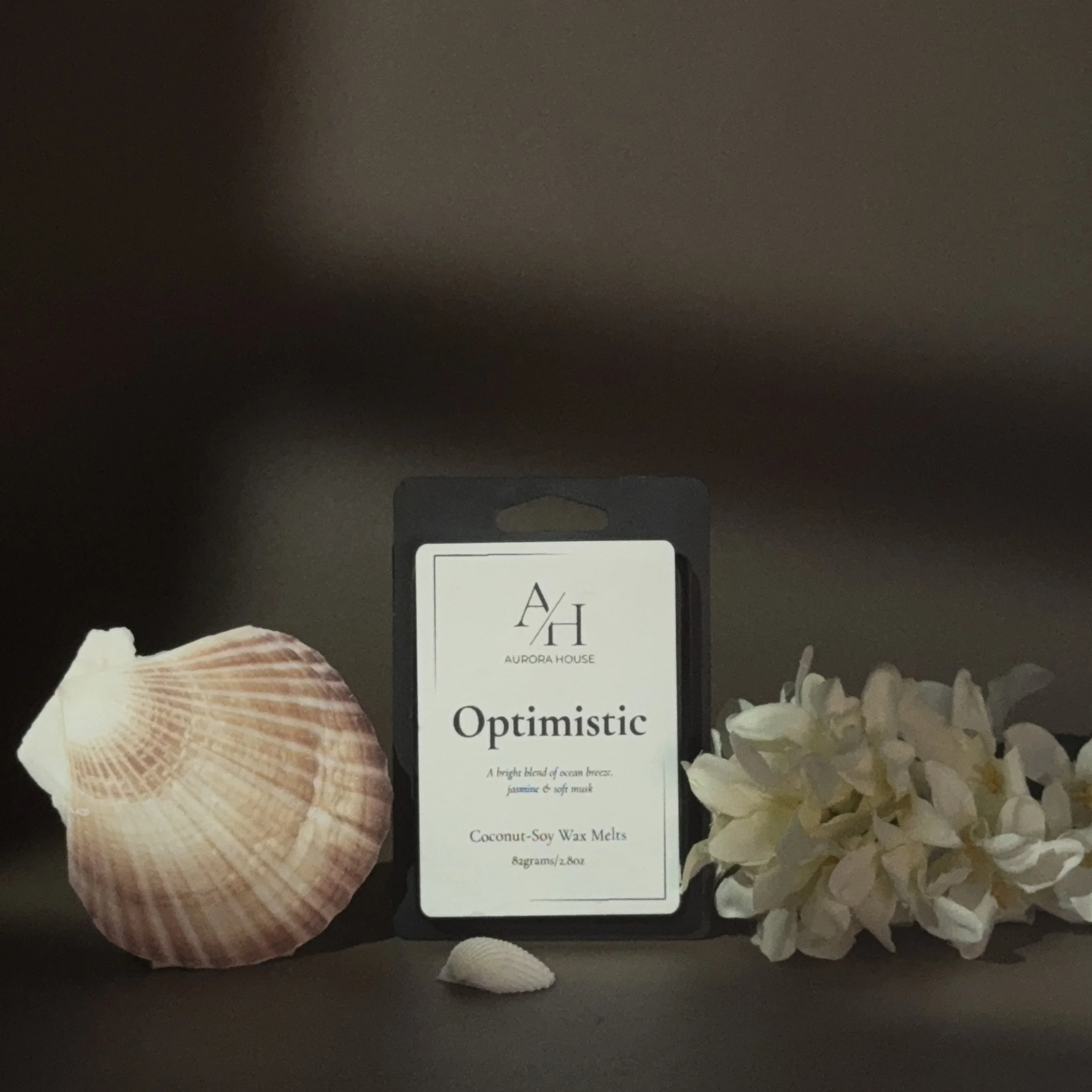 Package of Aurora House 'Optimistic' coconut-soy wax melts, 82 grams, with a seashell on the left and white flowers on the right on a gray surface.