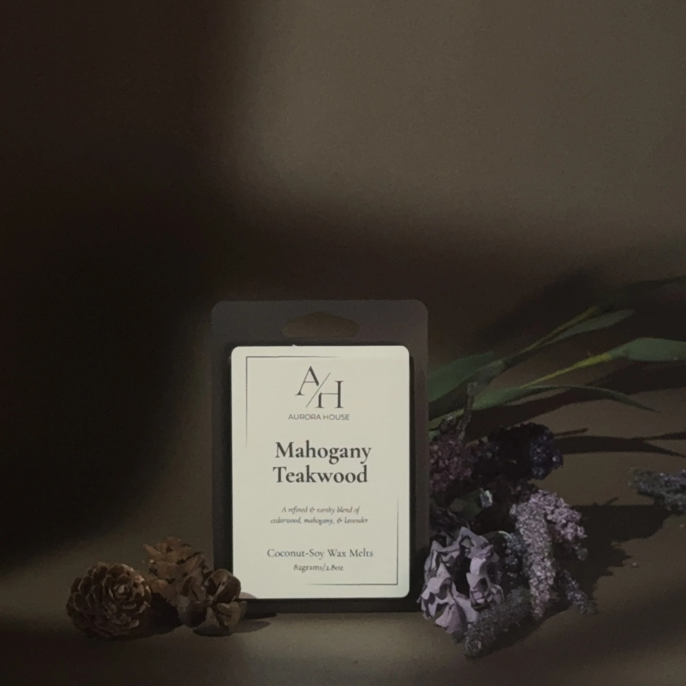 A black and white package of Auroras House Mahogany Teakwood coconut-soy wax melts on a wooden surface, with purple flowers and pinecones nearby.