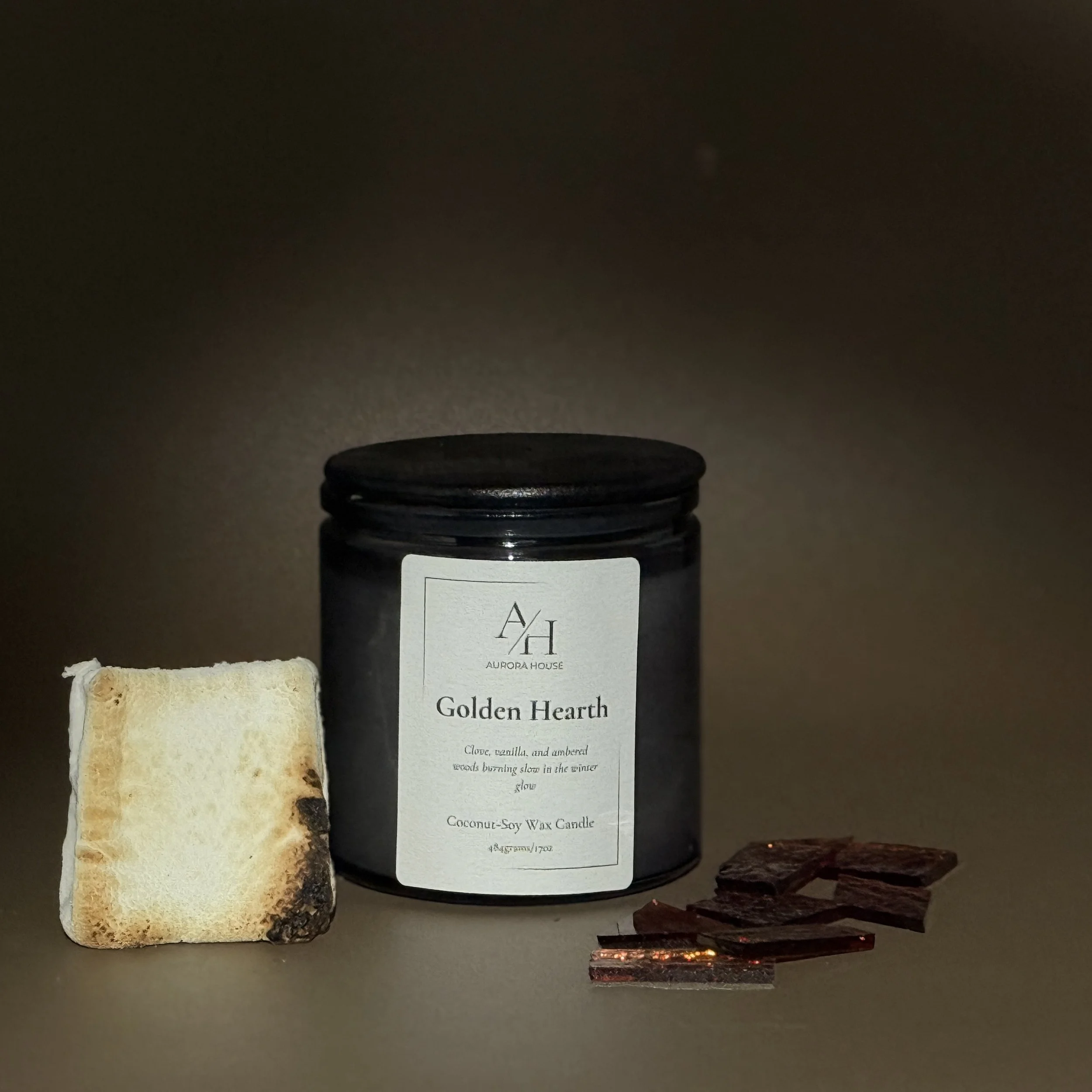 A black jar candle labeled "Golden Hearth" by Aurora House, with a toasted marshmallow on the left and small amber glass pieces on the right, all set against a neutral background.