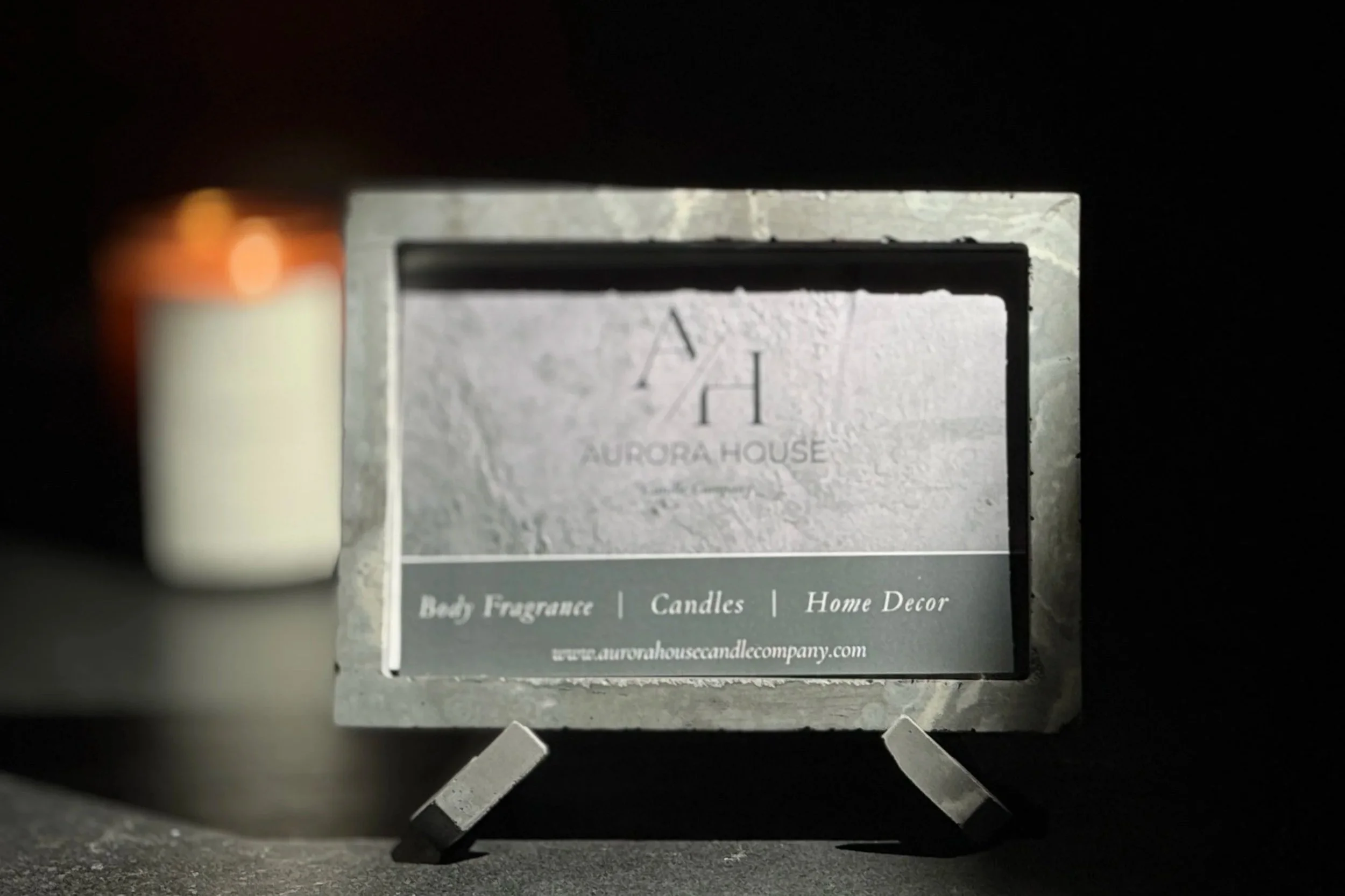 A small candle in a glass container with a white wax candle and a light-colored label. Behind it, there is a sign for Aurora House Candle Company, mentioning their products such as body fragrance, candles, and home decor.