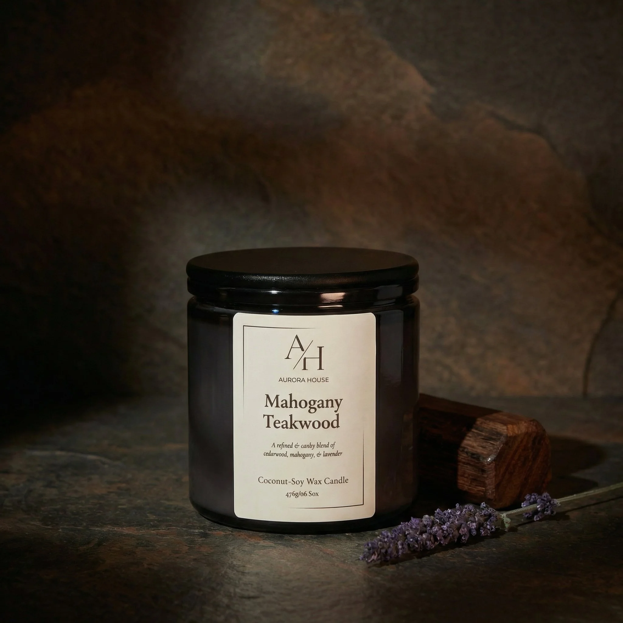 A black jar candle labeled 'Mahogany Teakwood' from Aurora House, with lavender and pinecones around it on a gray surface, against a beige background.