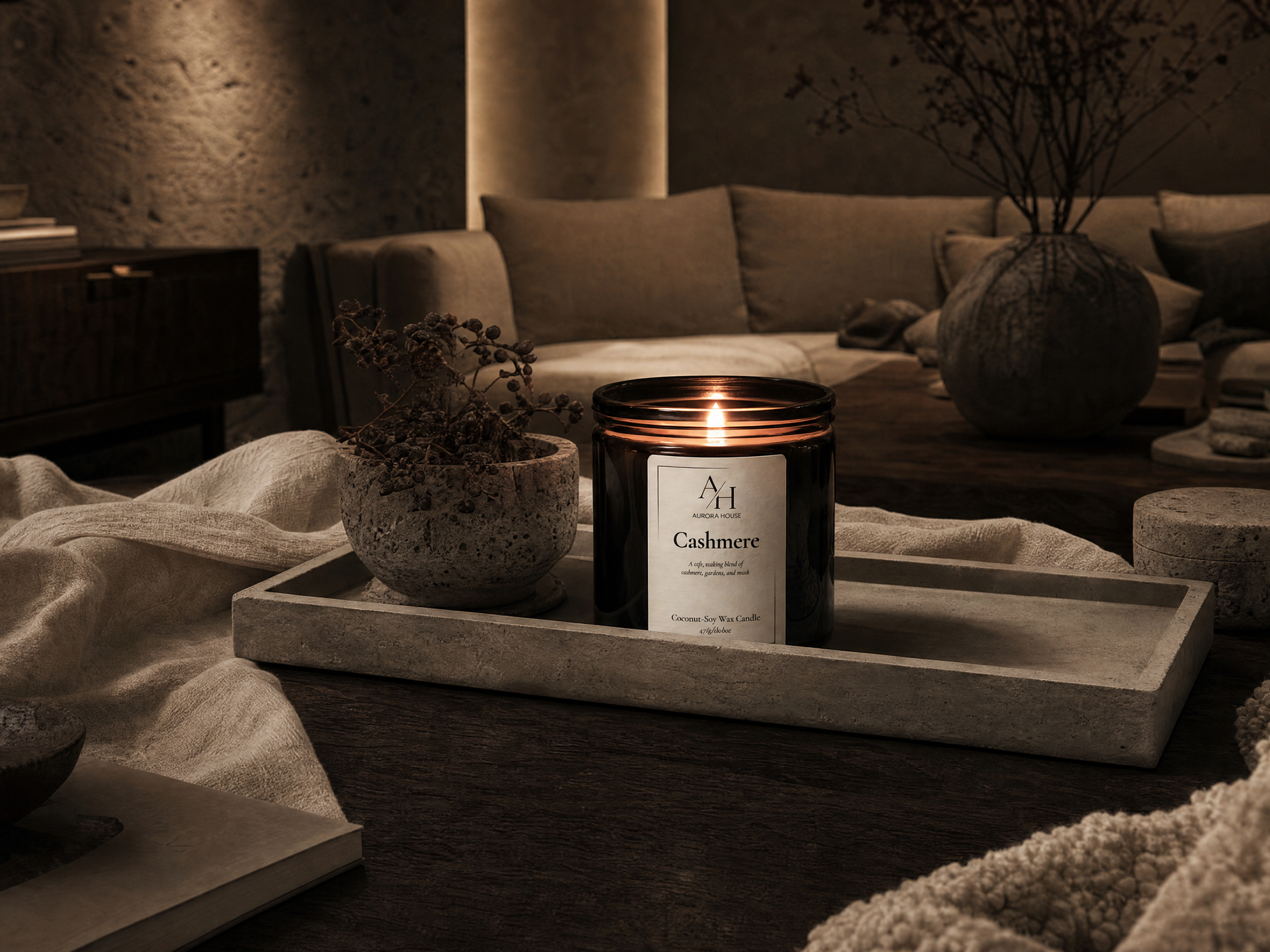 A lit candle on a tray with a label reading 'Cashmere' inside a cozy living room with beige couches, a large vase with branches, and textured walls.