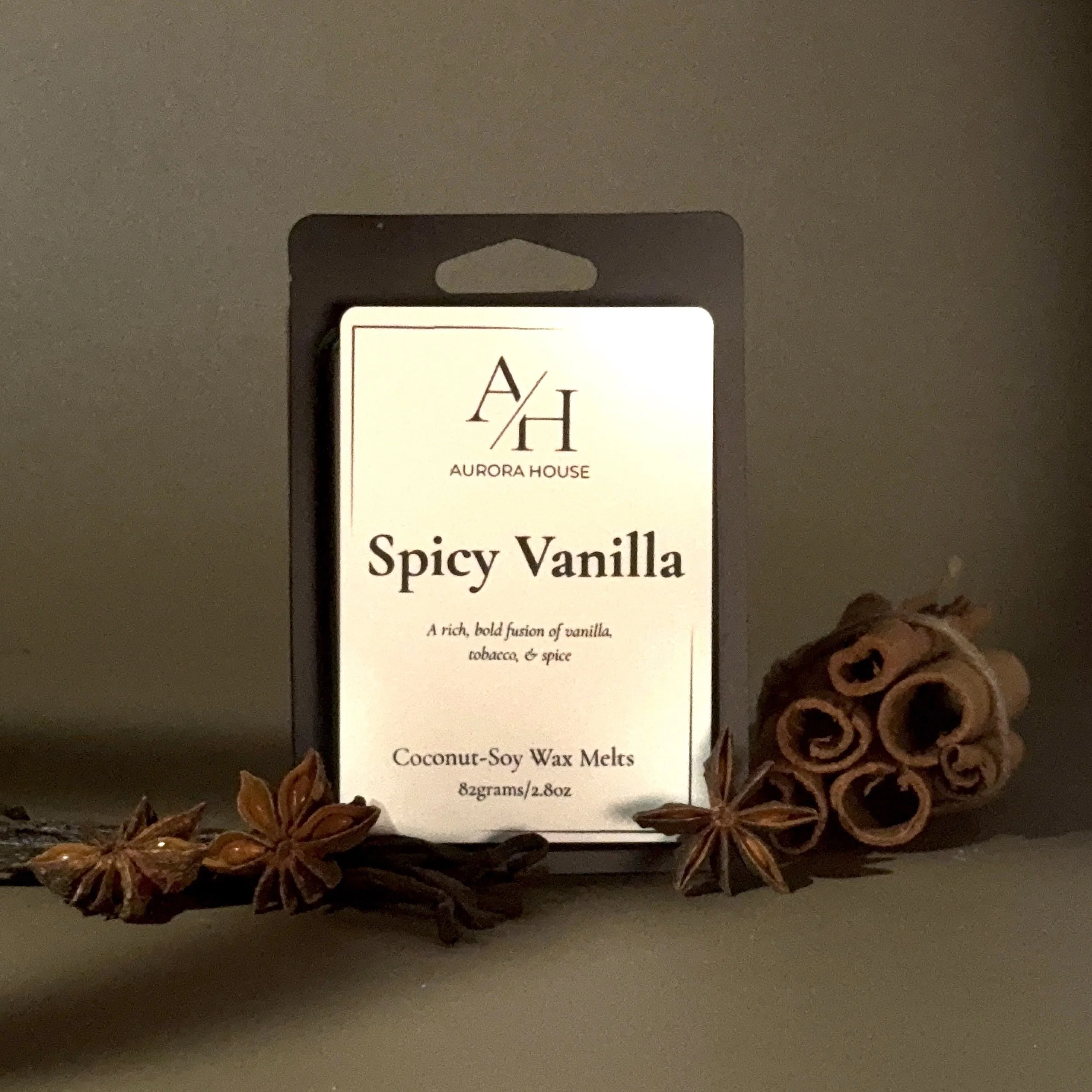 A package of Aurora House spicy vanilla coconut-soy wax melts on a wooden surface, with cinnamon sticks to the right and star anise to the left, against a plain wall background.