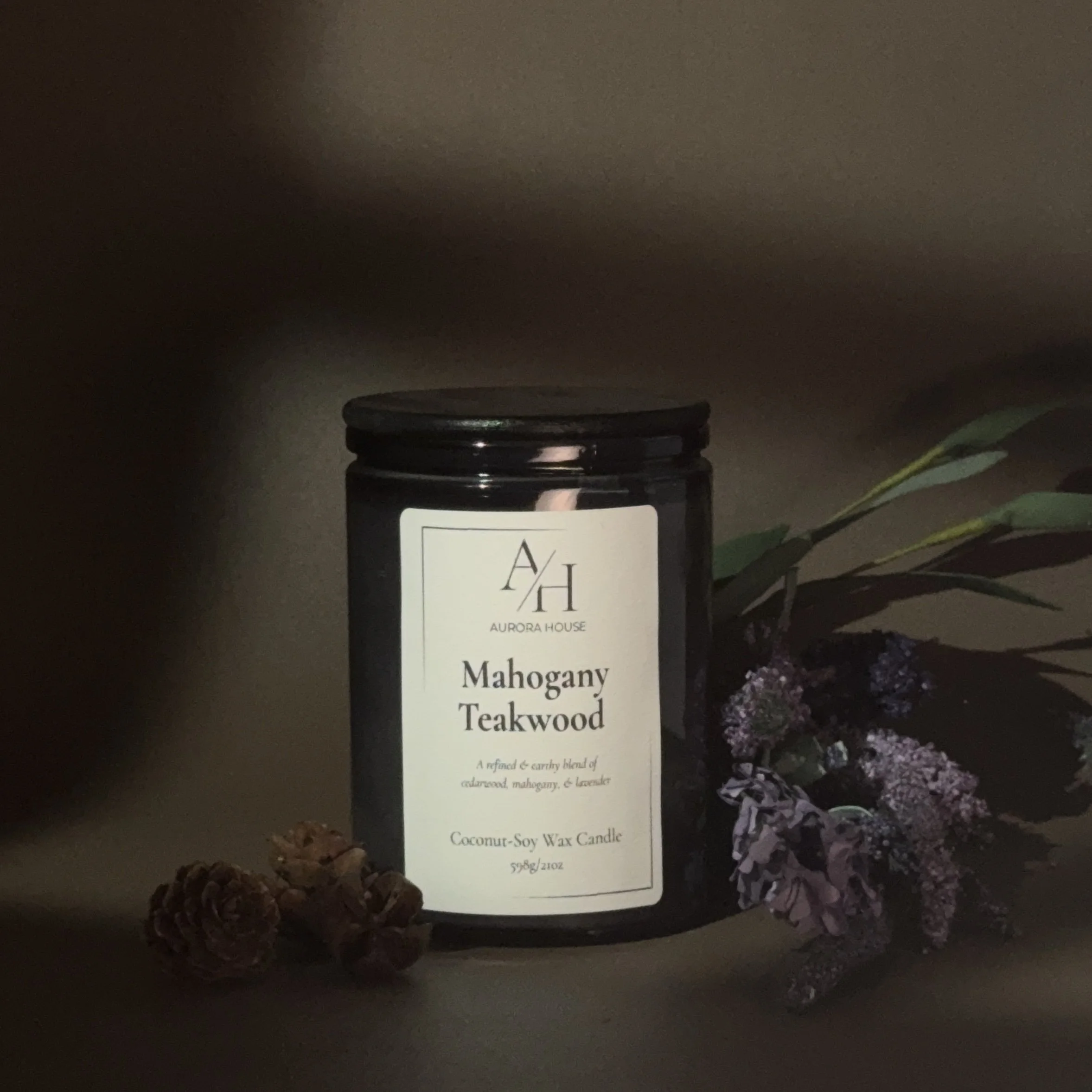 Black container with white label featuring Aurora House Mahogany Teakwood candle, placed on a wooden surface with pinecones and lavender flowers nearby.