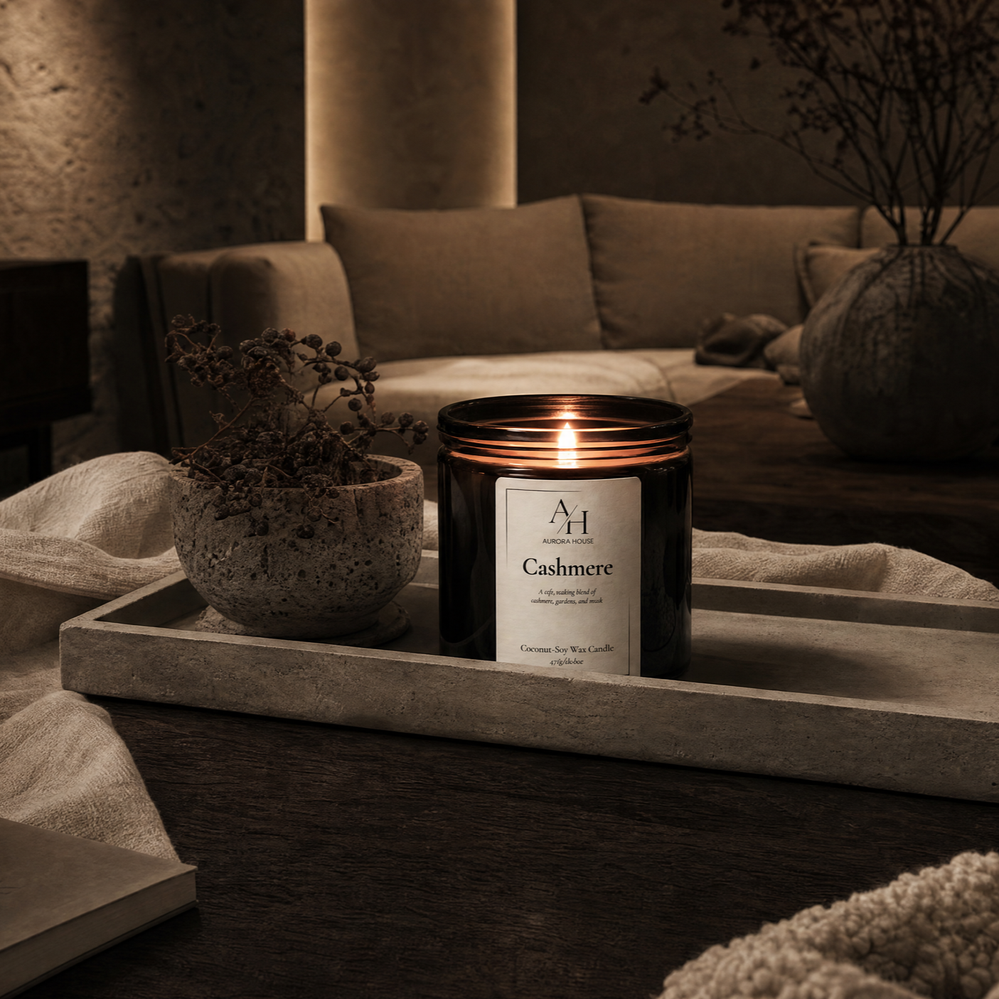 A lit black candle labeled 'Cashmere' on a wooden tray, with a textured ceramic pot with dried branches, in a cozy, warmly lit living room with beige sofa and dark textured wall.