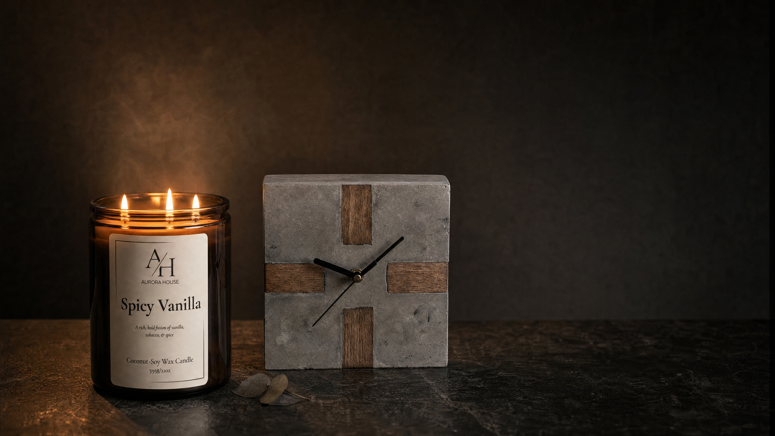 A lit candle labeled 'Spicy Vanilla' next to a small modern clock with a concrete and wood design on a dark surface, against a dark background.