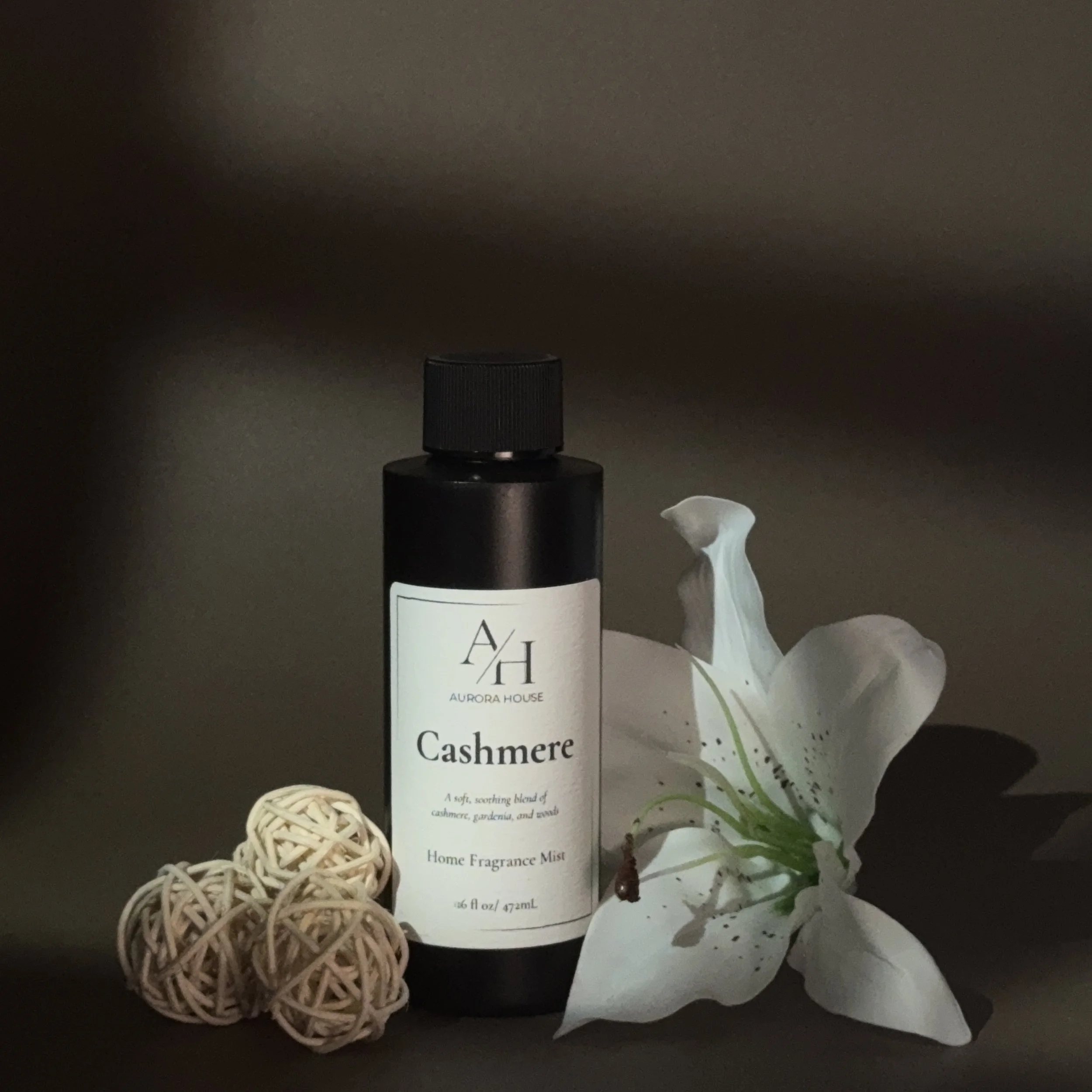 A black bottle of Aurora House Cashmere home fragrance mist, with a white lily flower and beige decorative balls on a gray surface against a beige wall.