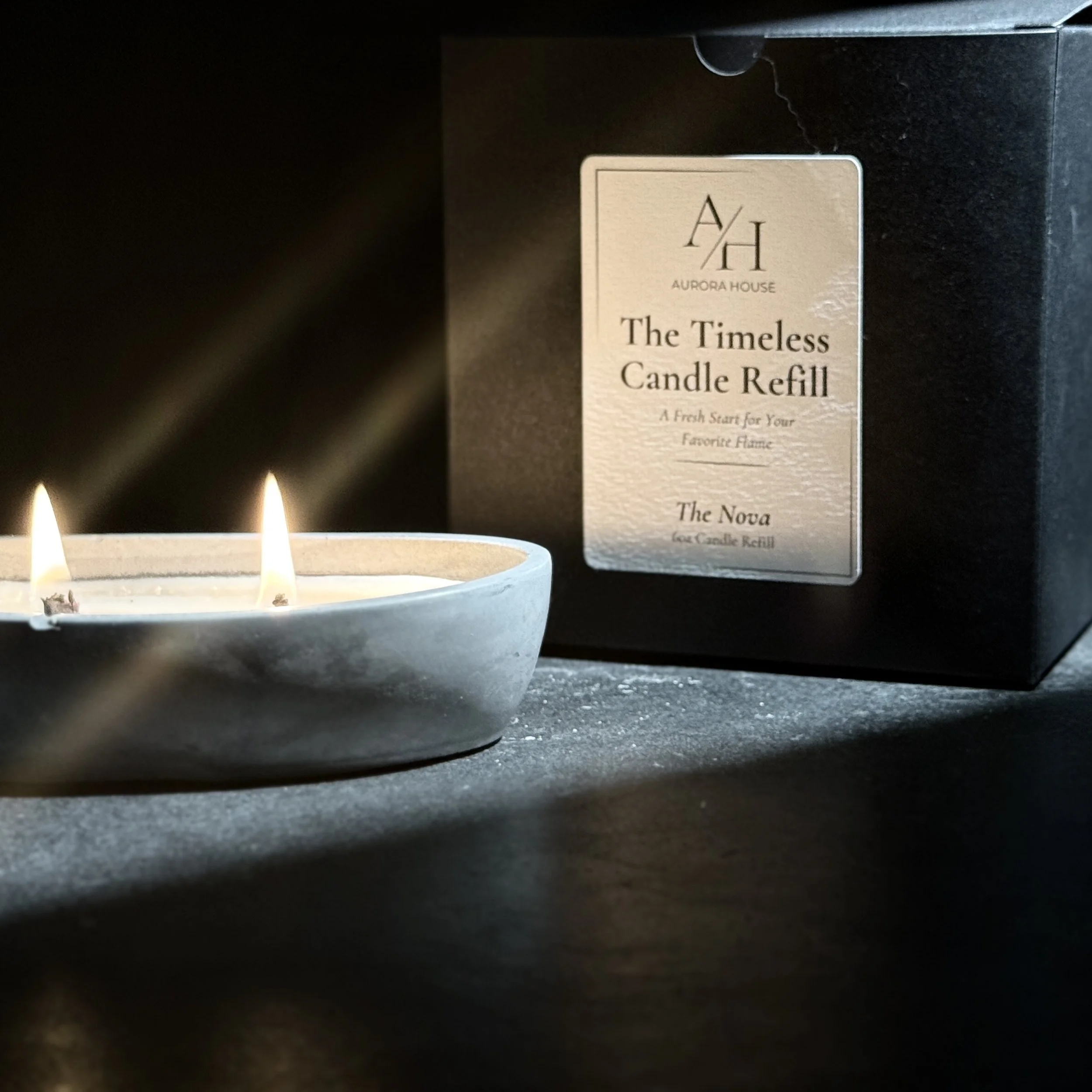 A lit candle in a small bowl-shaped holder and a black box labeled 'Aurora House The Timeless Candle Refill' with the subtitle 'A Fresh Start for Your Favorite Flame' and the scent name 'The Nova'.
