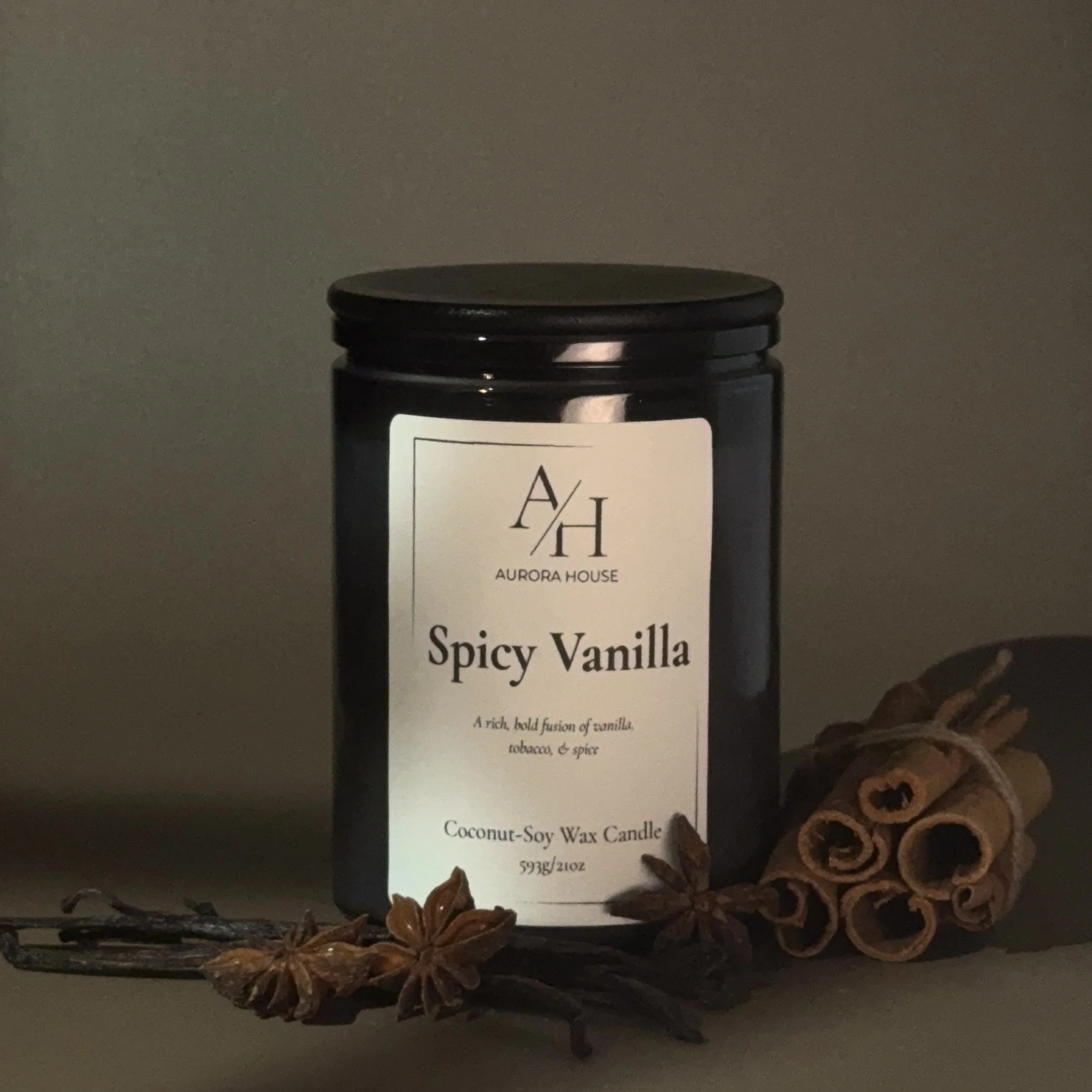 A black jar labeled Aurora House Spicy Vanilla coconut-soy wax candle, with star anise and cinnamon sticks in front of it against a gray textured background.