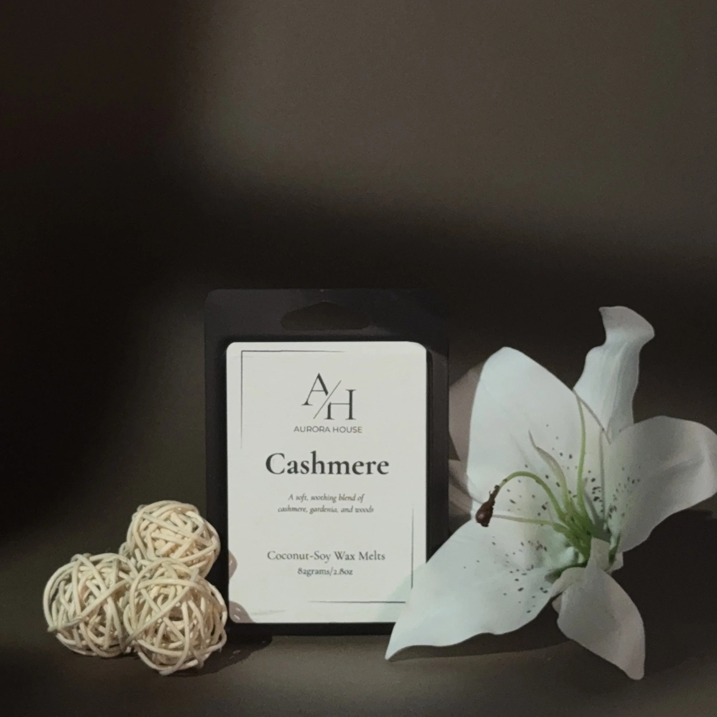 A package of Aurora House Cashmere Coconut-Soy Wax Melts standing upright on a surface, with a white flower on the right and decorative ball on the left.