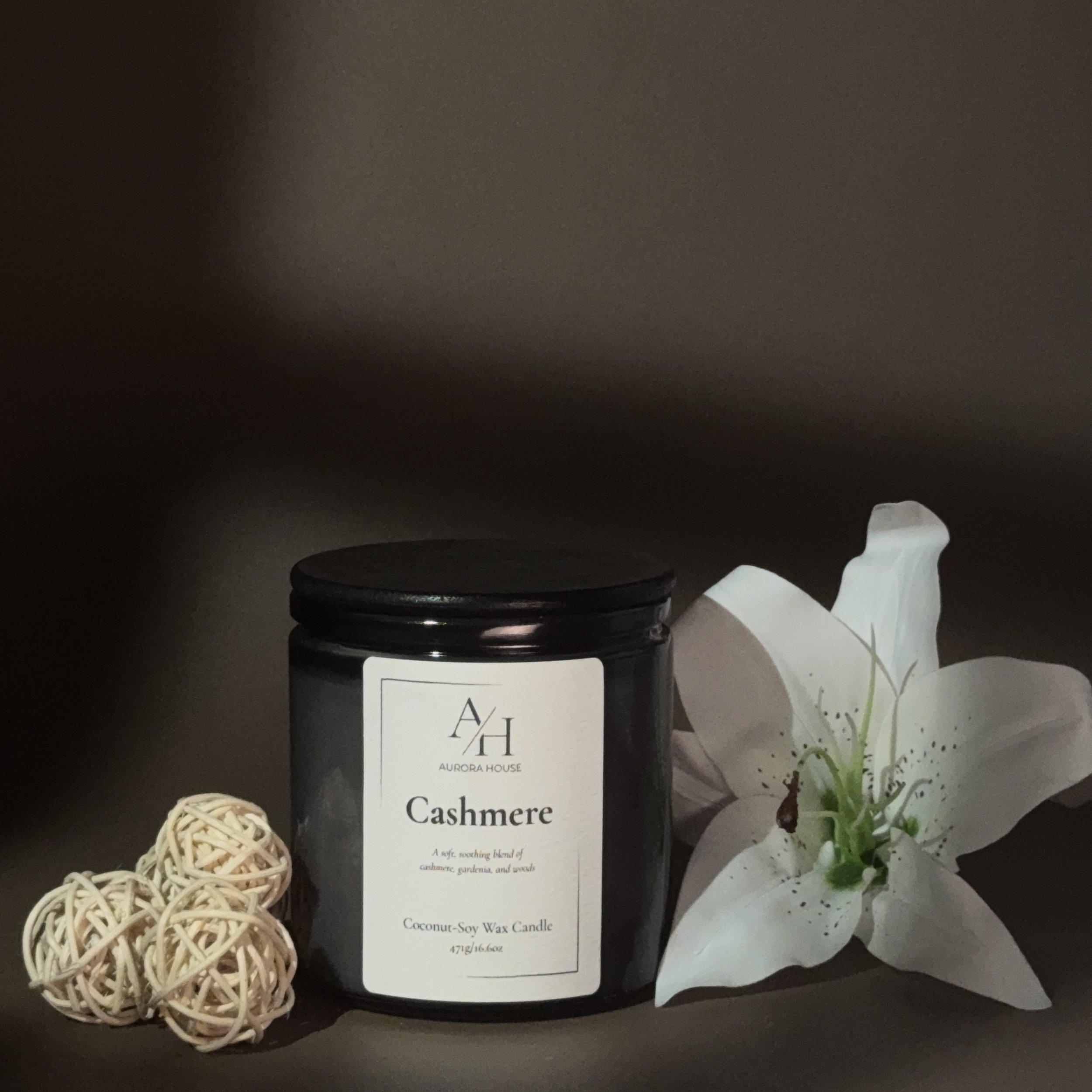 A black jar of Aurora House Cashmere coconut soy wax candle with a white label, surrounded by white decorative flowers and rattan ball on a gray surface.