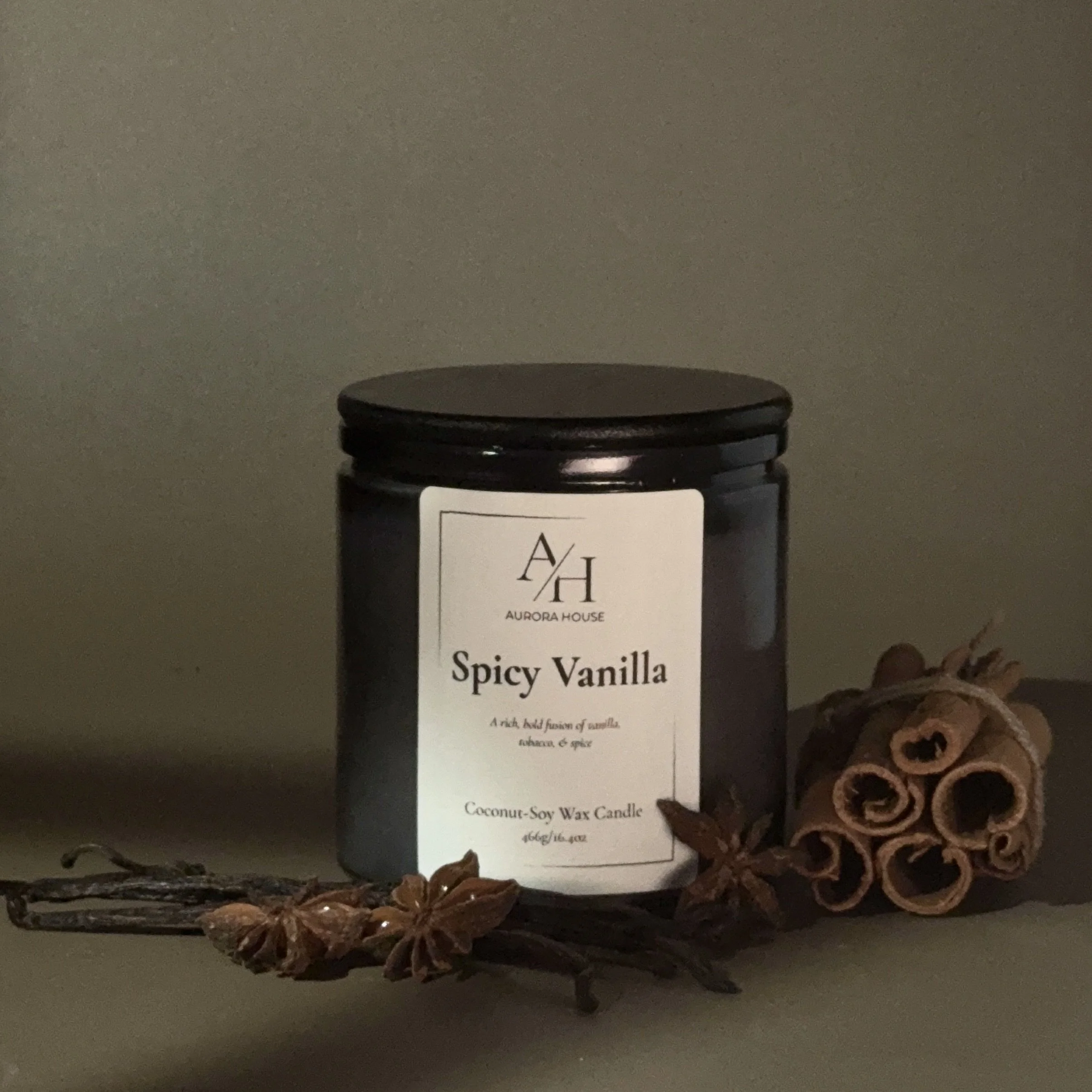 A black candle jar labeled 'Aurora House Spicy Vanilla' with cinnamon sticks and star anise nearby on a light gray surface.