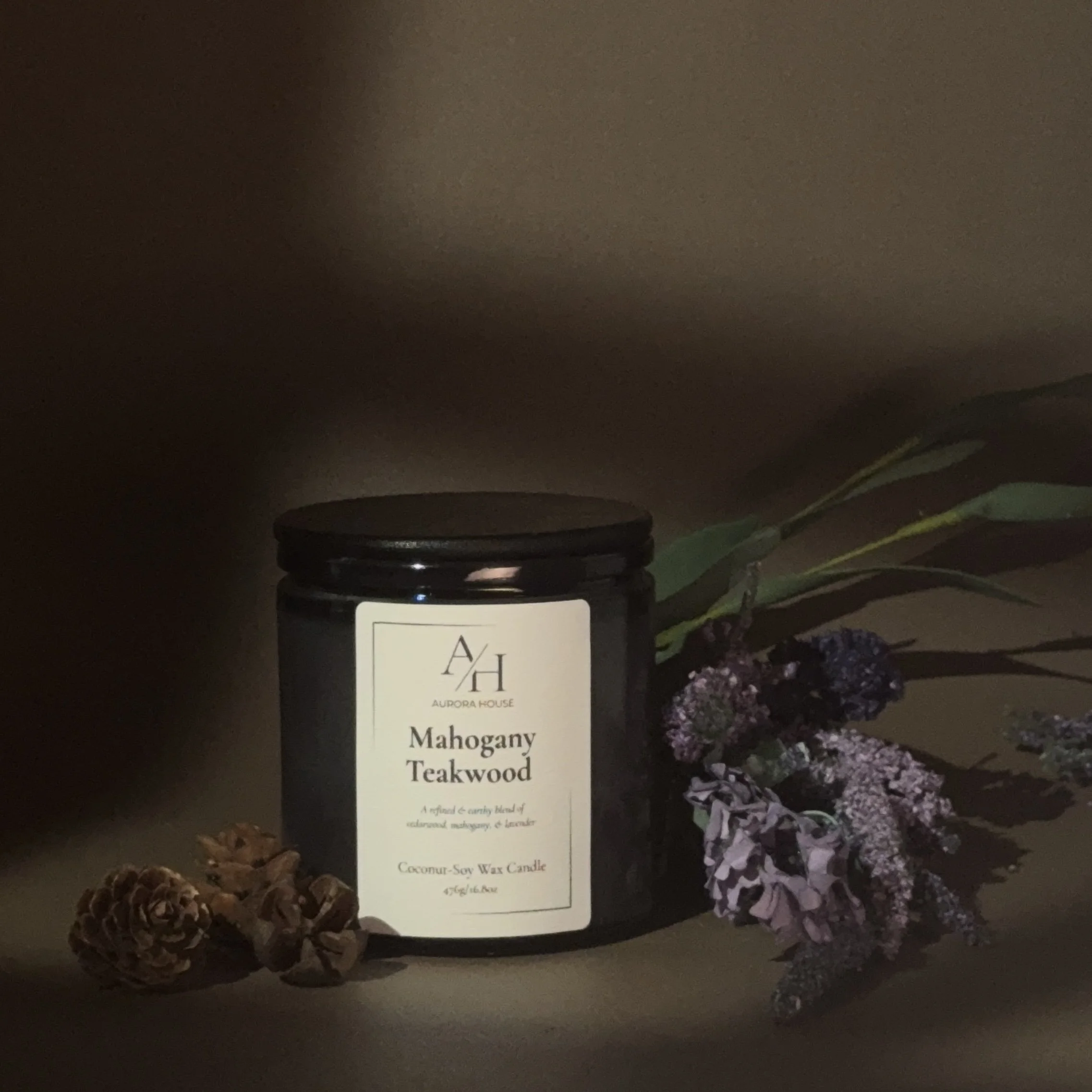 A black jar candle labeled 'Mahogany Teakwood' from Aurora House, with lavender and pinecones around it on a gray surface, against a beige background.