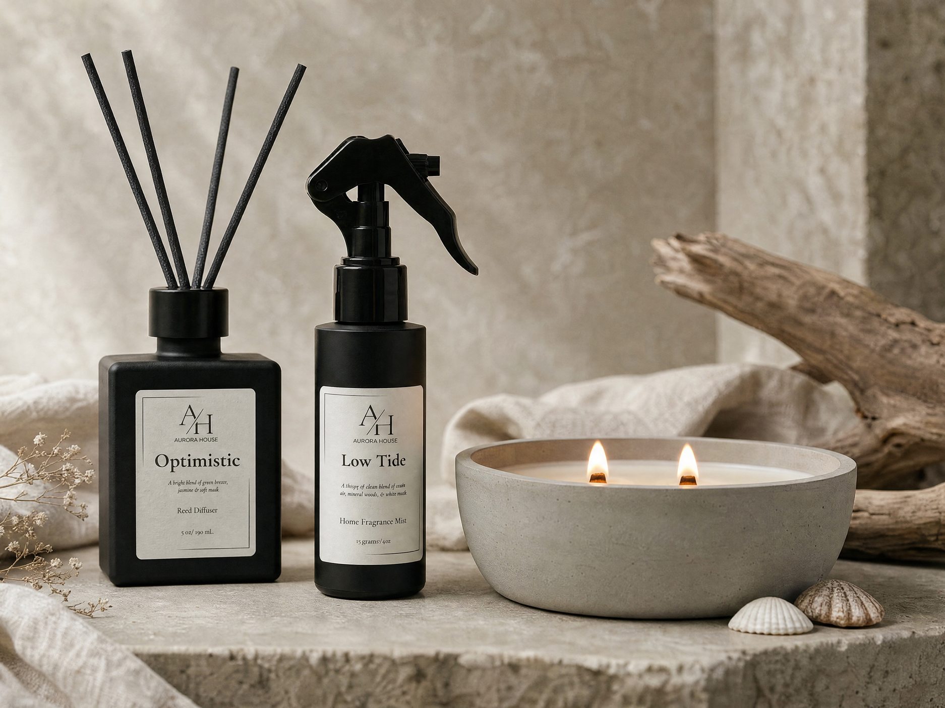 Two black bottles of home fragrance mist labeled "Optimistic" and "Low Tide" from Aurora House, with white labels and black caps, placed beside a lit white ceramic candle holder, with seashells, dried flowers, a piece of driftwood, and a beige linen cloth on a textured surface in the background.