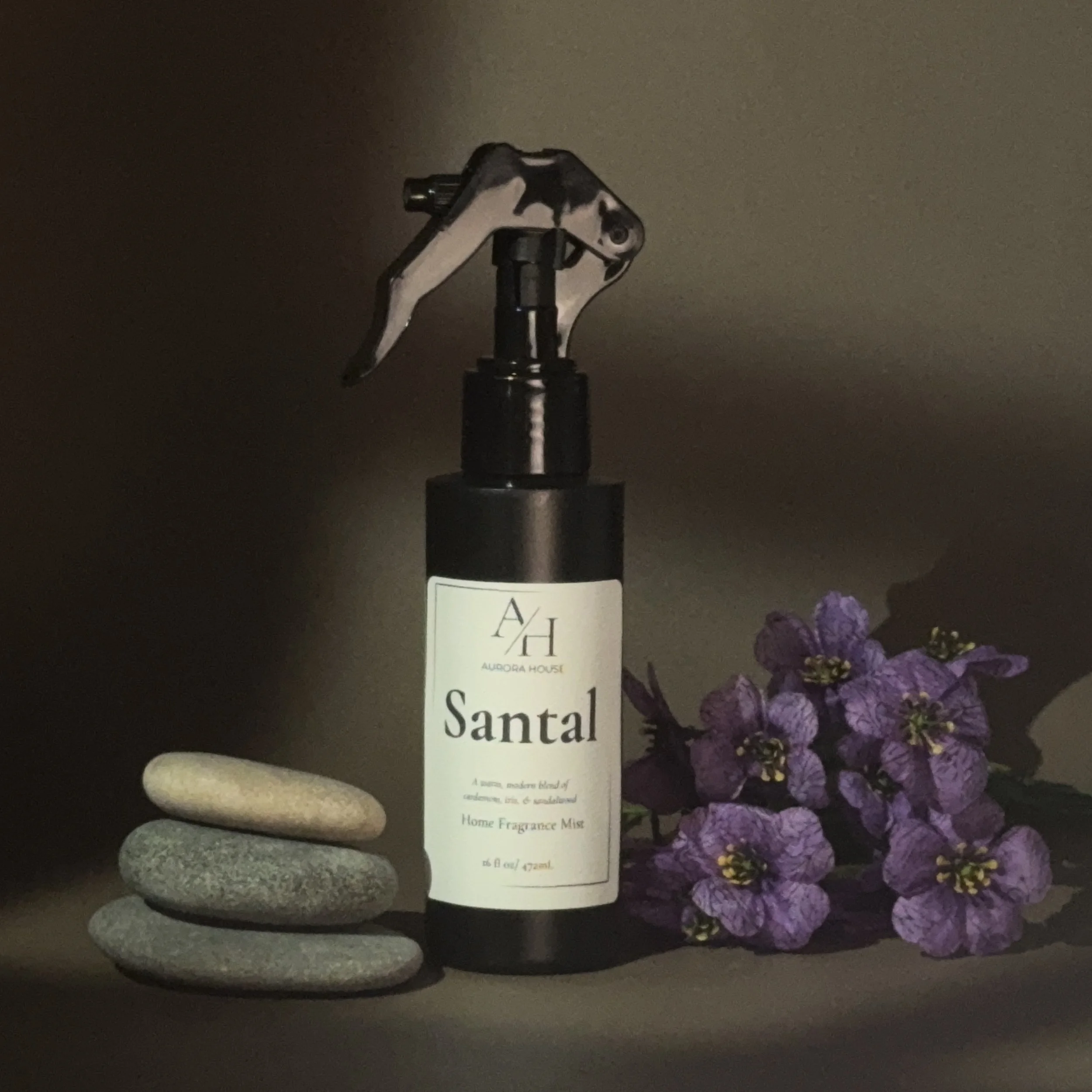 A black spray bottle labeled 'Santal' by Aurora House, surrounded by three stacked smooth stones on the left and purple flowers on the right, against a neutral background.