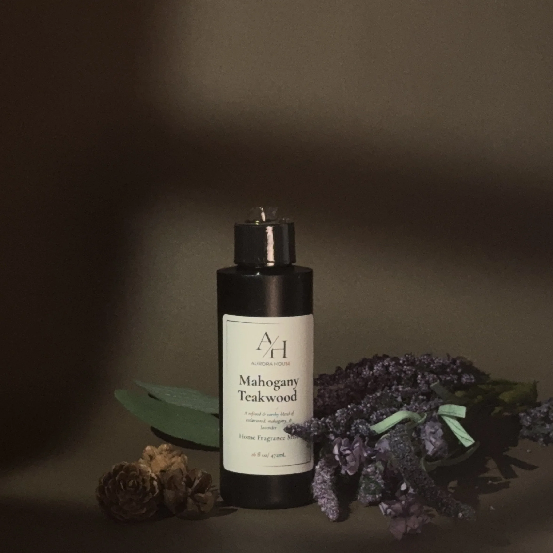 A black bottle labeled 'Mahogany Teakwood' home fragrance mist from Aurora House, with lavender and purple flowers and pinecones arranged on a gray surface.