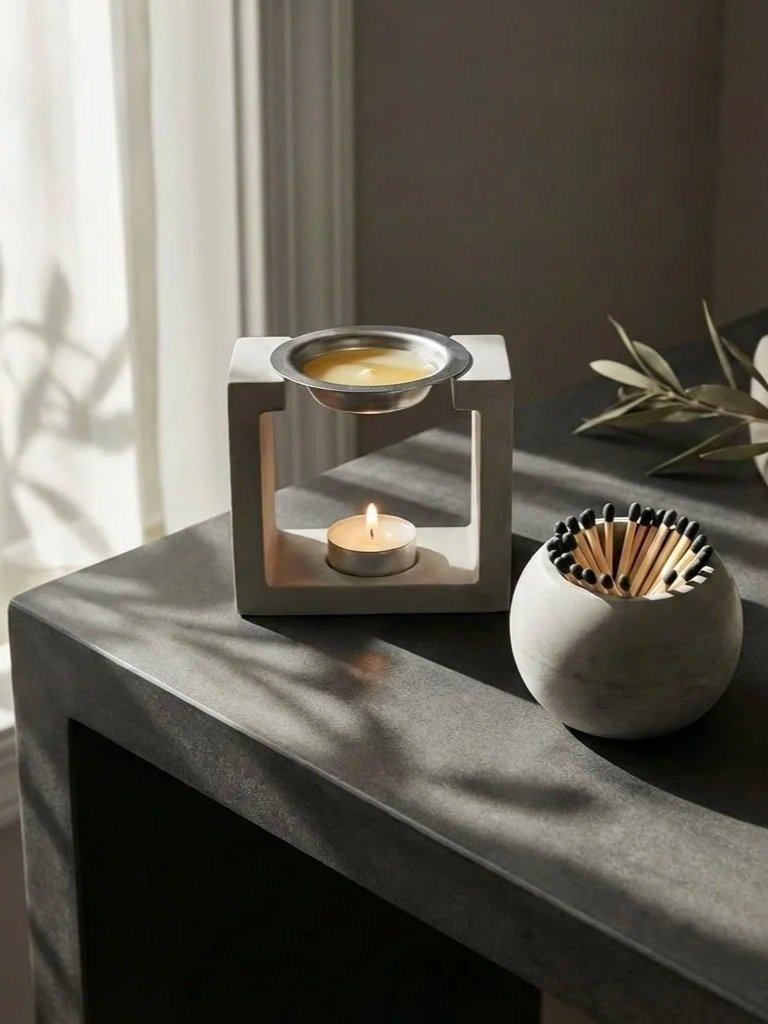 A modern candle holder with a lit tea light candle, a small dish with melted wax, and a round container filled with black-tipped matchsticks on a dark gray table, with sunlight casting shadows.