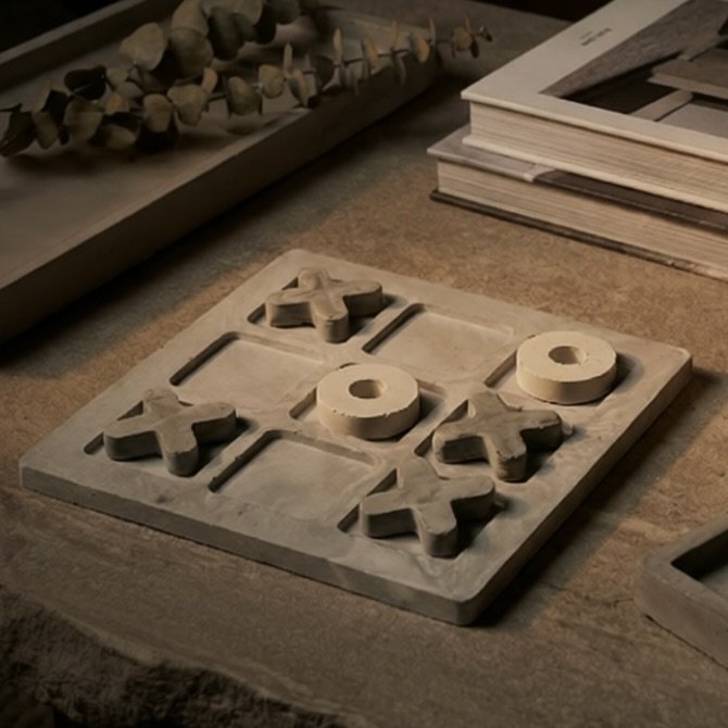 A wooden tic-tac-toe game with X and O pieces, placed on a wooden table with books and decorative items in the background.