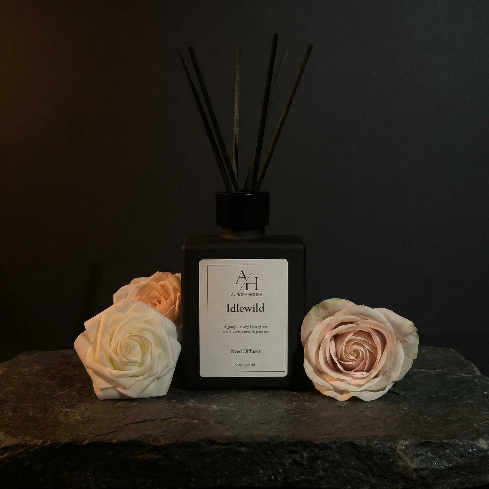 A black reed diffuser bottle labeled 'Idlewild' from Aurora House, with black reeds, a white artificial rose, and green foliage on a brown surface against a brown background.