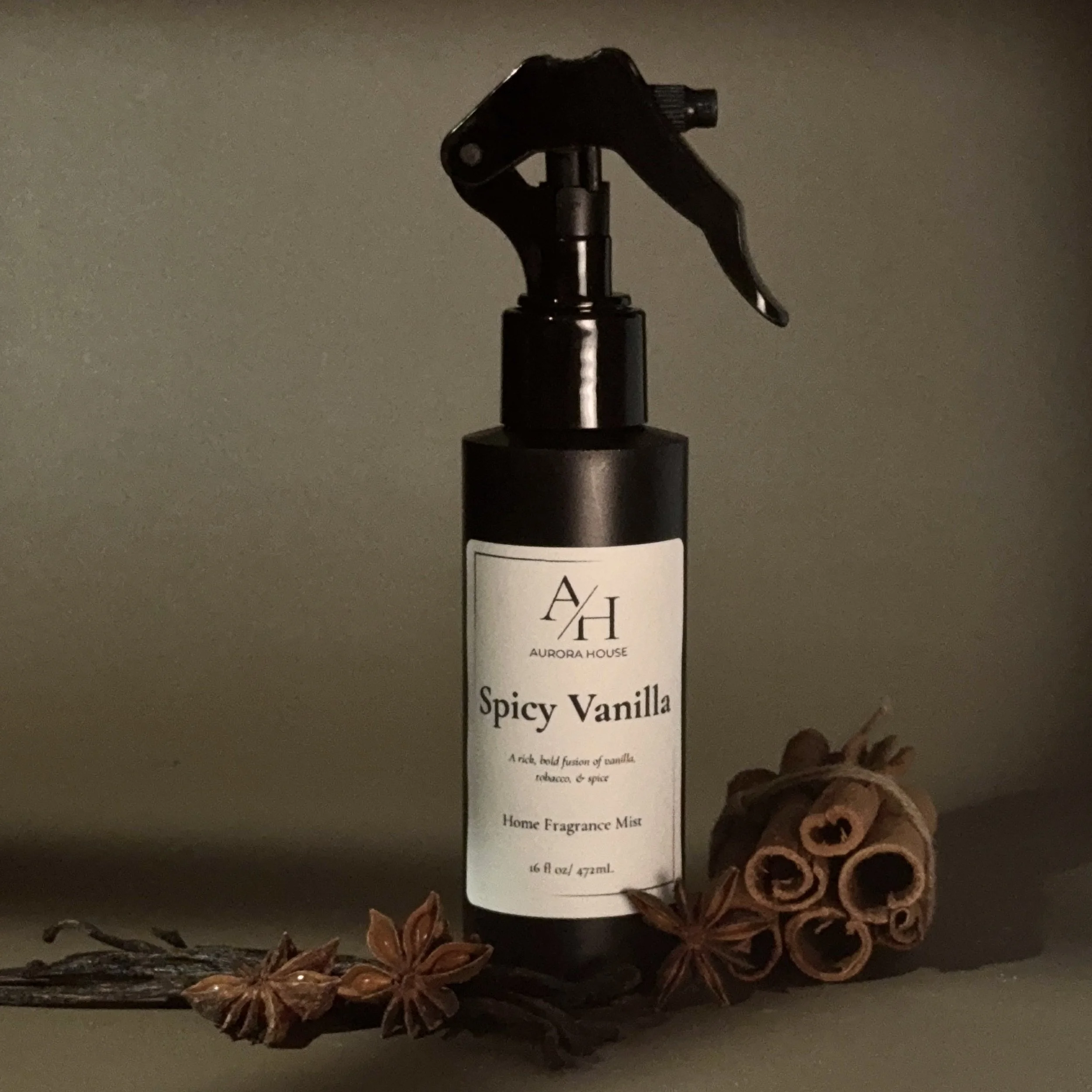 A black spray bottle labeled Aurora House Spicy Vanilla Home Fragrance Mist, with cinnamon sticks and star anise in front of it on a gray surface against a beige background.