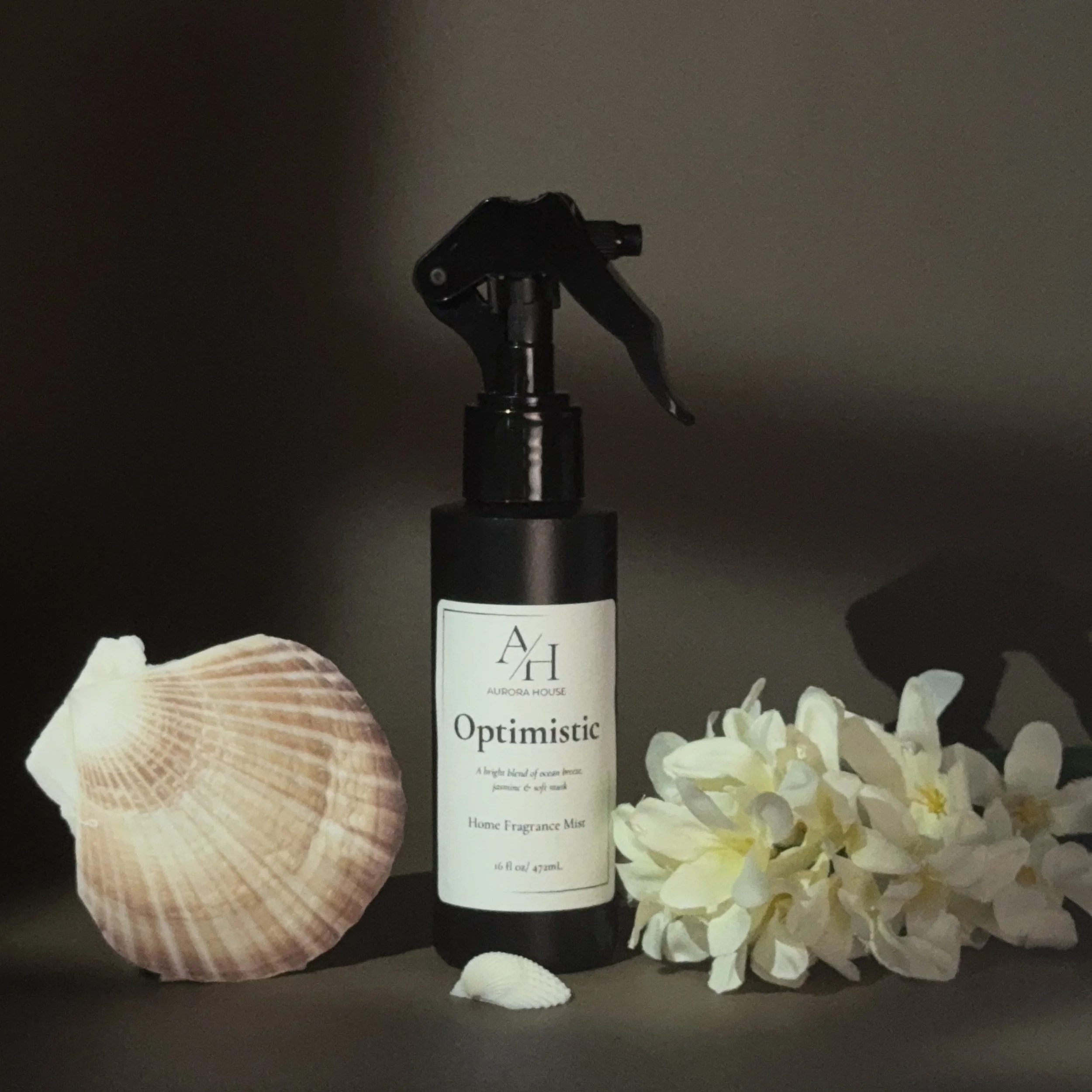 A black spray bottle labeled 'Optimistic' with a scent description, flanked by a pink seashell on the left and white flowers on the right, set against a gray background.