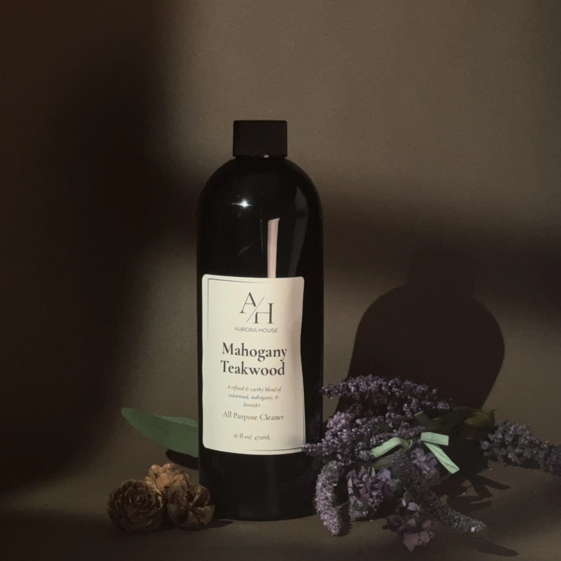 A black bottle of Aurora House Mahogany Teakwood all purpose cleaner with a white label featuring black text, placed on a wooden surface with pinecones and purple flowers around it.