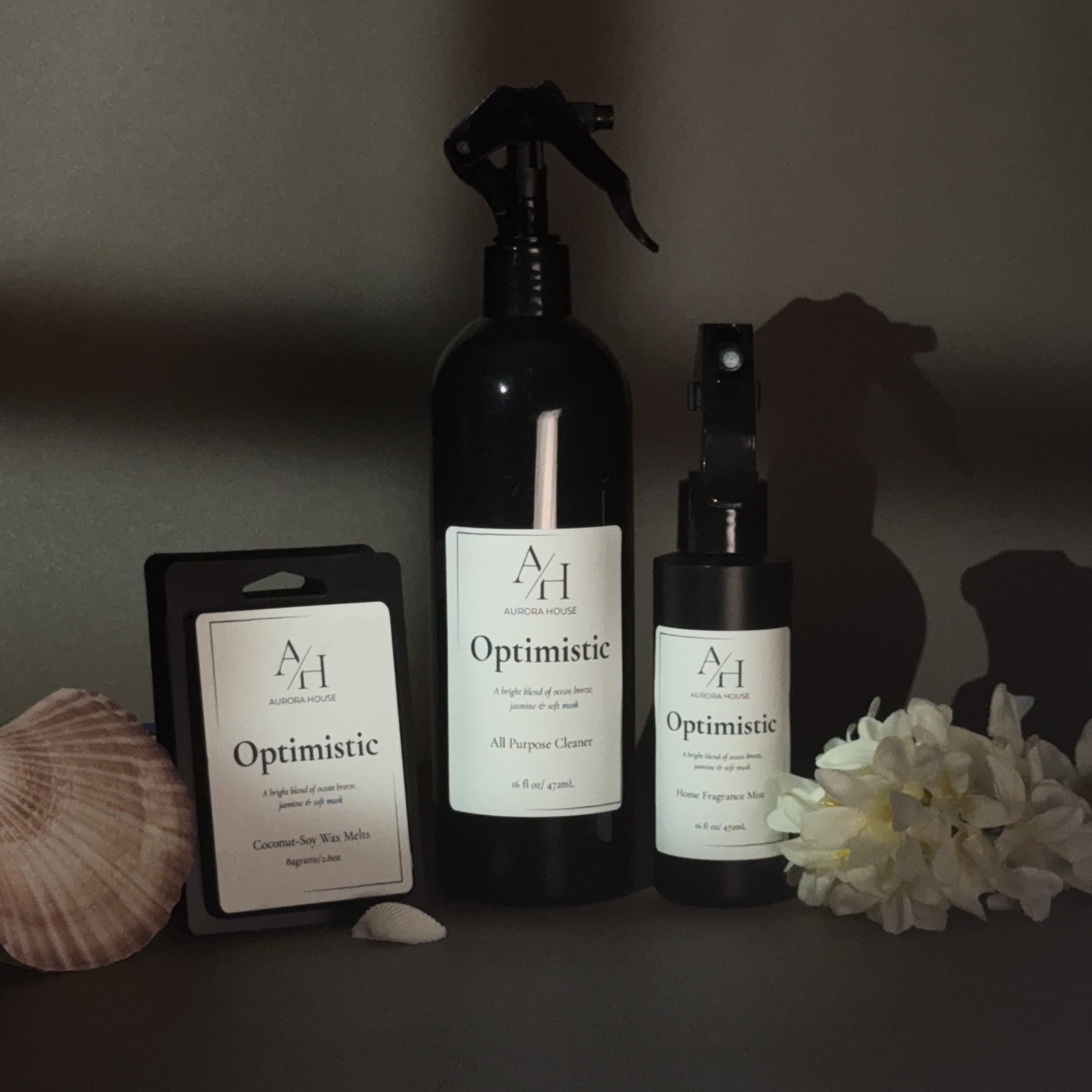 Collection of Aurora House products including an all-purpose cleaner in a spray bottle, a home fragrance mist in a pump bottle, and coconut-soy wax melts, with a seashell and white flowers beside them.