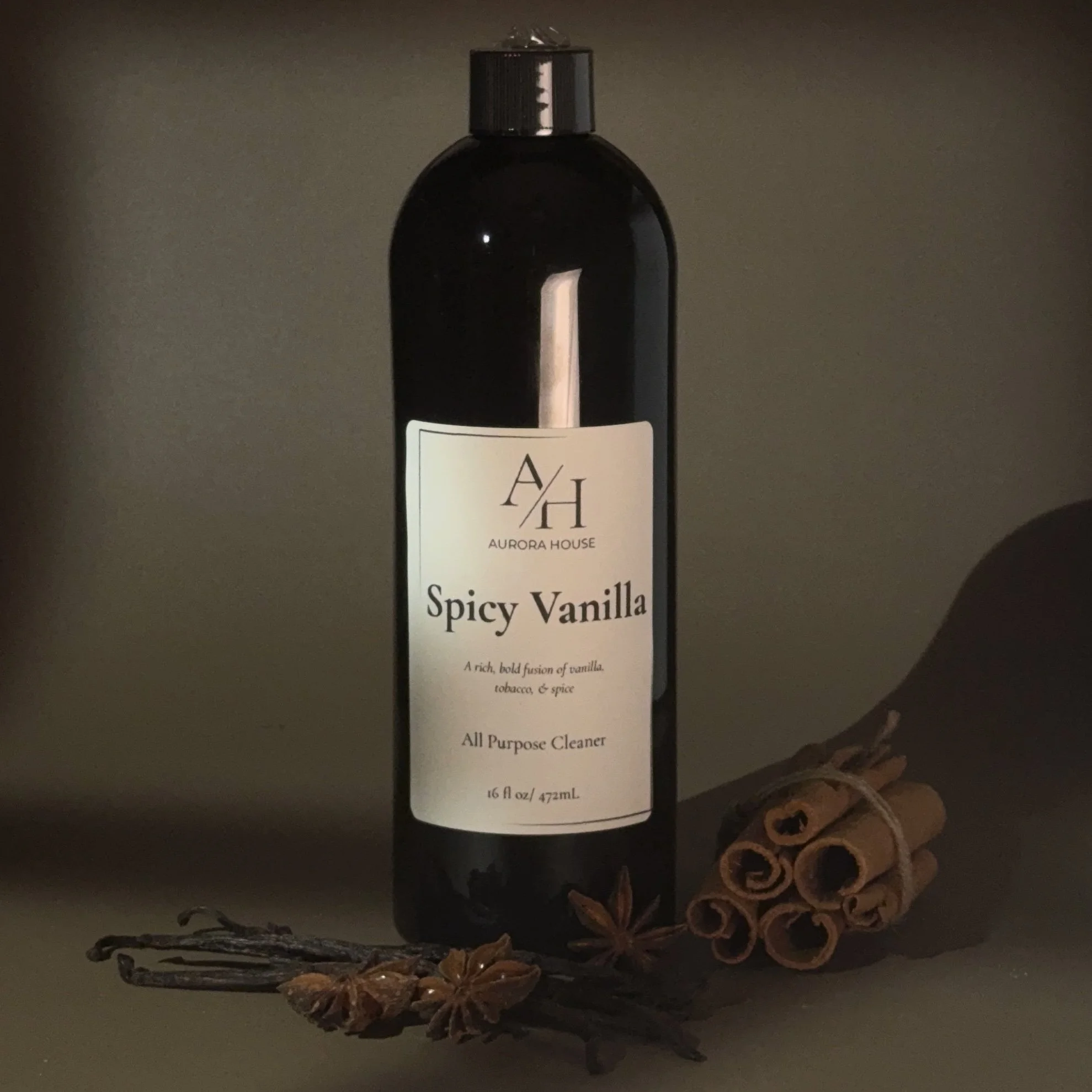 A black bottle of Aurora House Spicy Vanilla all-purpose cleaner with a white label, placed on a gray surface with cinnamon sticks and star anise in front.
