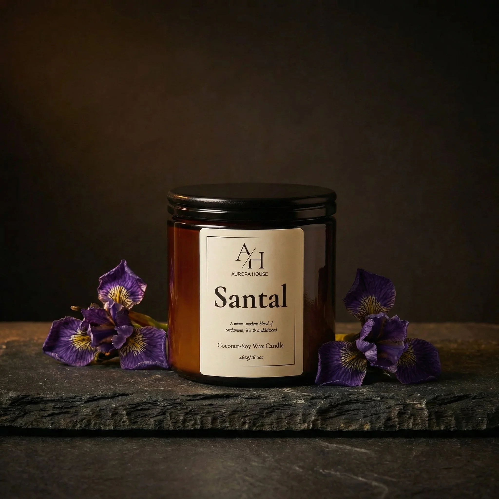 A black candle jar labeled 'Santal' by Aurora House, with purple flowers and stacked gray stones beside it on a light-colored surface.