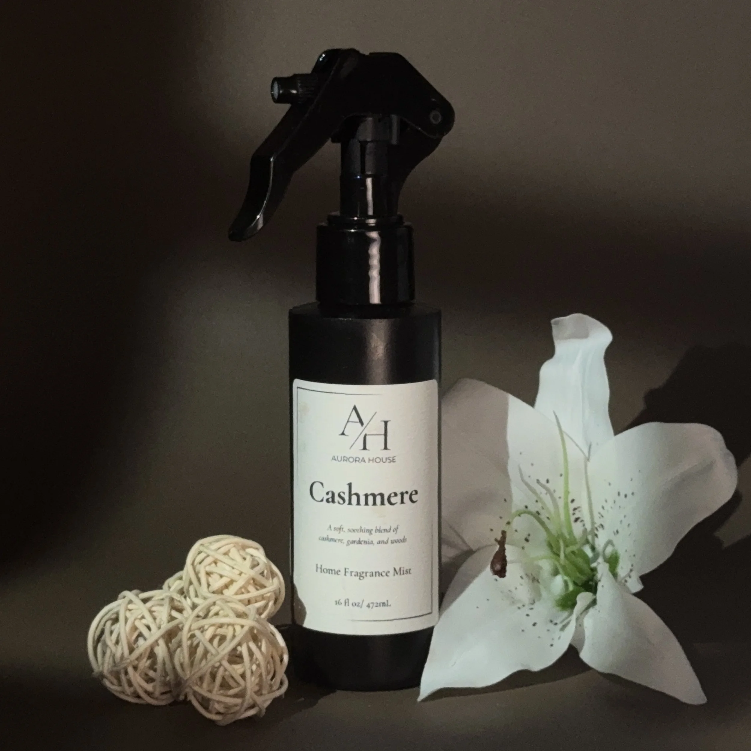 A spray bottle labeled Aurora House Cashmere Home Fragrance Mist, three decorative rattan balls, and a white lily flower, arranged on a flat surface against a neutral background.