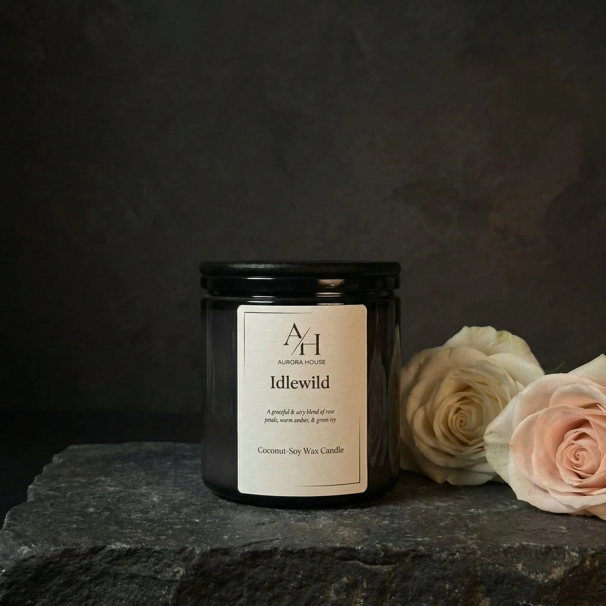A black jar of Aurora Rose candle labeled Idlewild with white roses and green leaves around it on a gray surface against a neutral background.