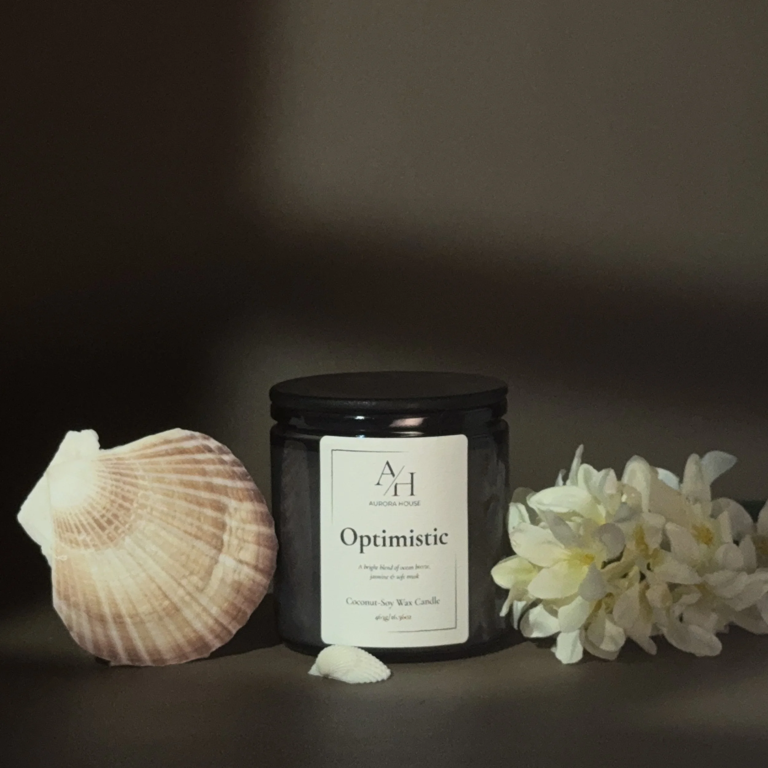 A black candle jar labeled 'Optimistic' from Aurora House, described as a coconut-soy wax candle with an optimistic, joyful, and peaceful scent, on a gray surface with a seashell on the left and white flowers on the right.