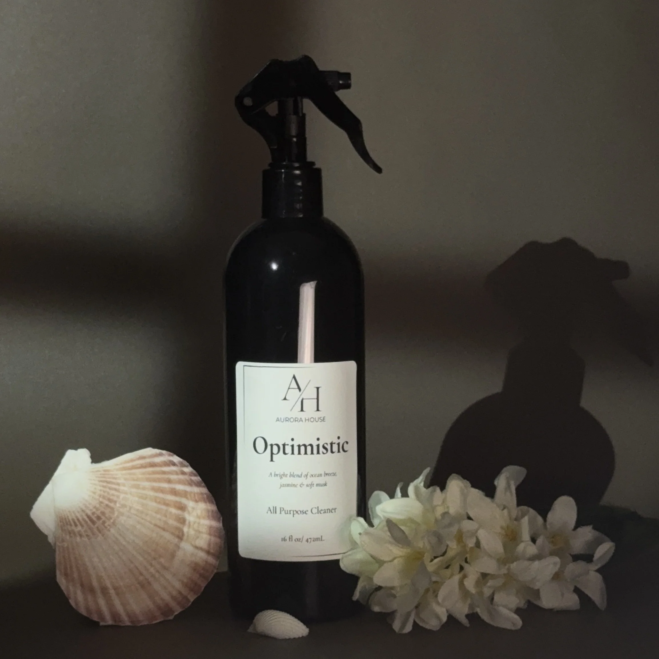 A black spray bottle labeled Aurora House Optimistic, surrounded by a seashell on the left and white flowers on the right, against a plain gray background.