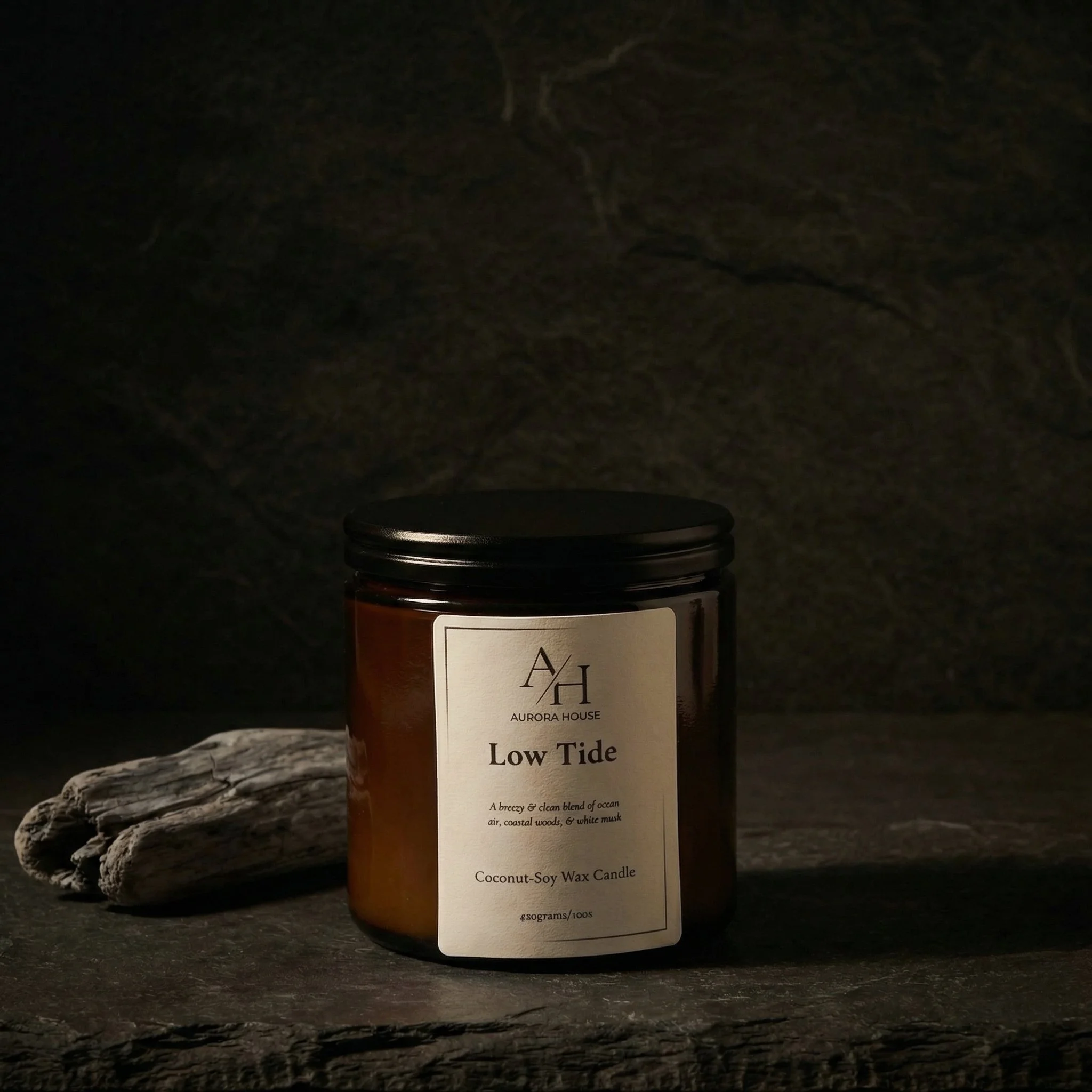 A black jar of Aurora House Low Tide Coconut-Soy Wax Candle on a light-colored surface with blue wax pieces and a dried plant branch nearby.