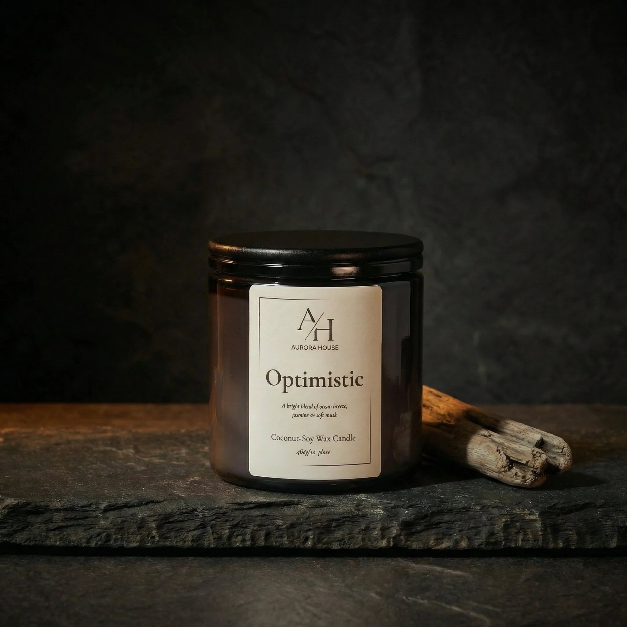 A black candle jar labeled 'Optimistic' from Aurora House, described as a coconut-soy wax candle with an optimistic, joyful, and peaceful scent, on a gray surface with a seashell on the left and white flowers on the right.