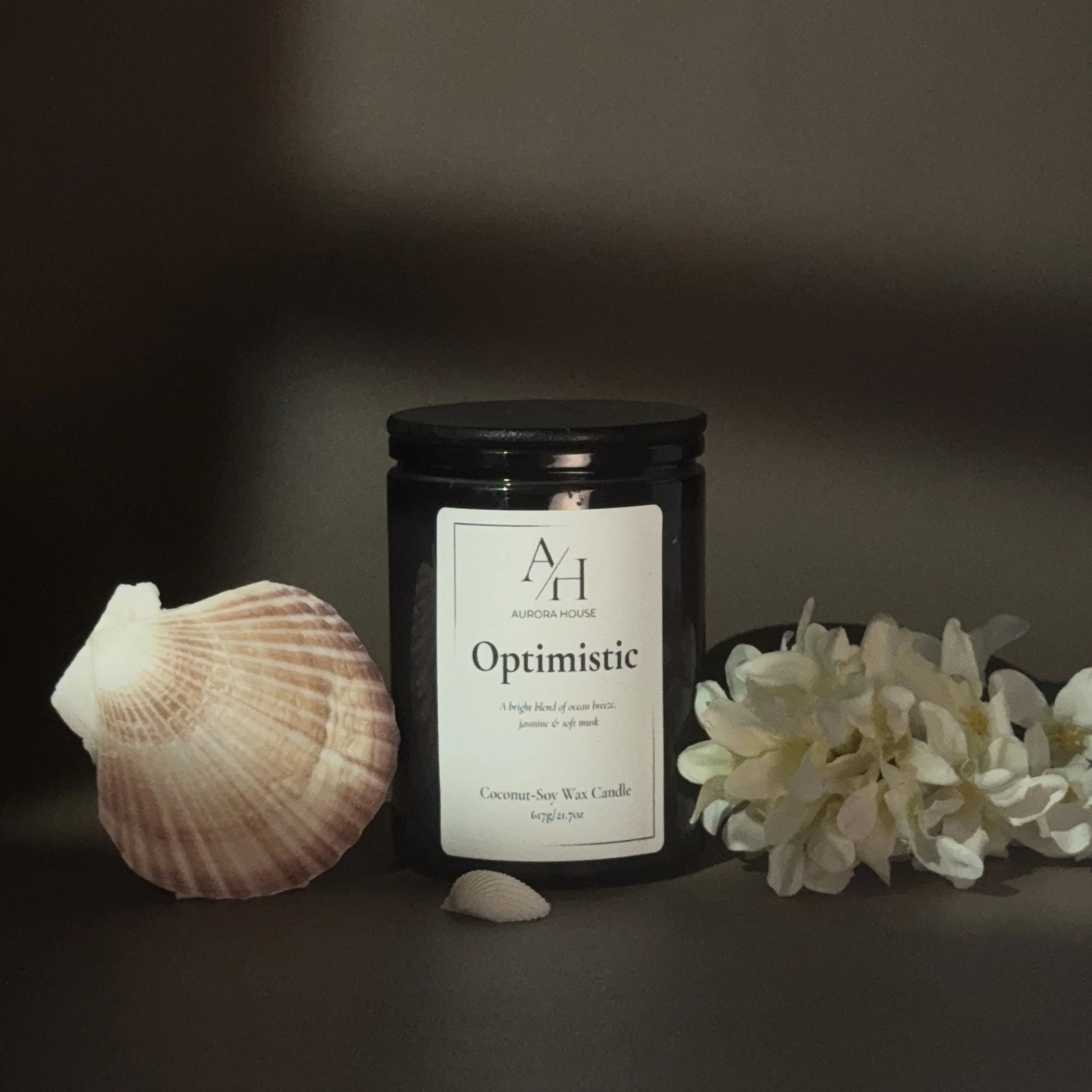 A black jar candle labeled Aurora House 'Optimistic' with coconut-soy wax, surrounded by a pink seashell on the left and white flowers on the right, on a gray surface against a neutral background.