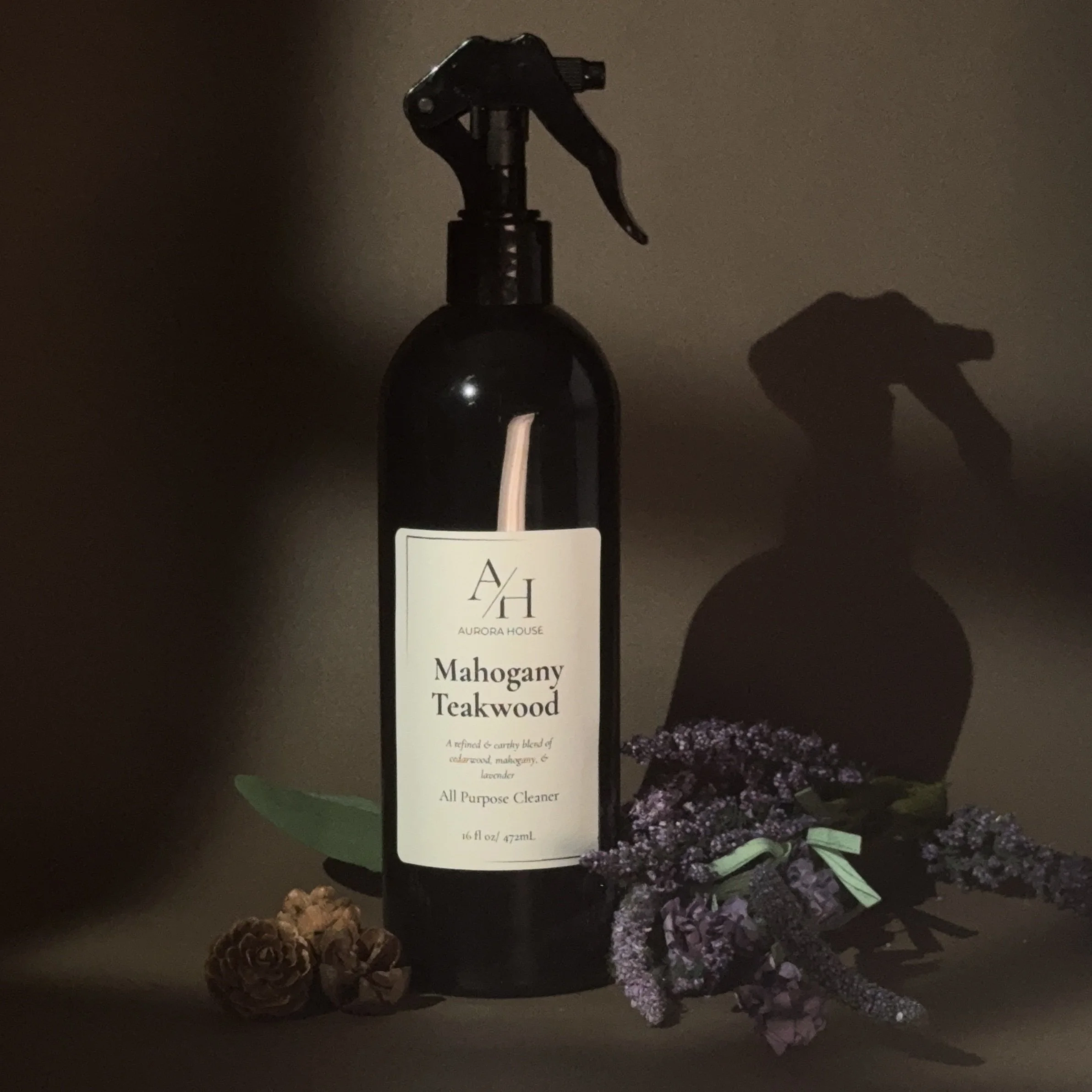 Black spray bottle labeled Aurora House Mahogany Teakwood all purpose cleaner with purple and green flower and pine cones on a wooden surface.