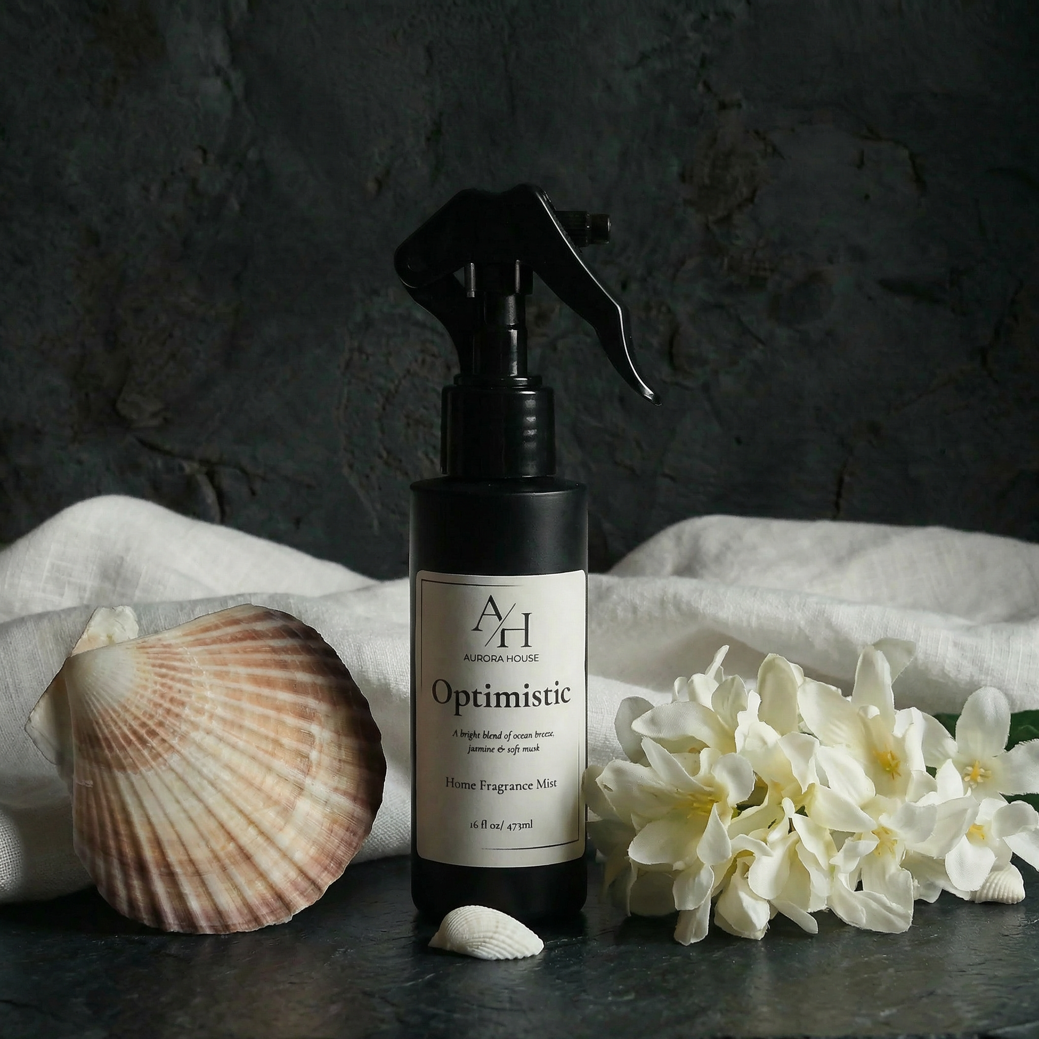 A black spray bottle labeled 'Optimistic' with a scent description, flanked by a pink seashell on the left and white flowers on the right, set against a gray background.