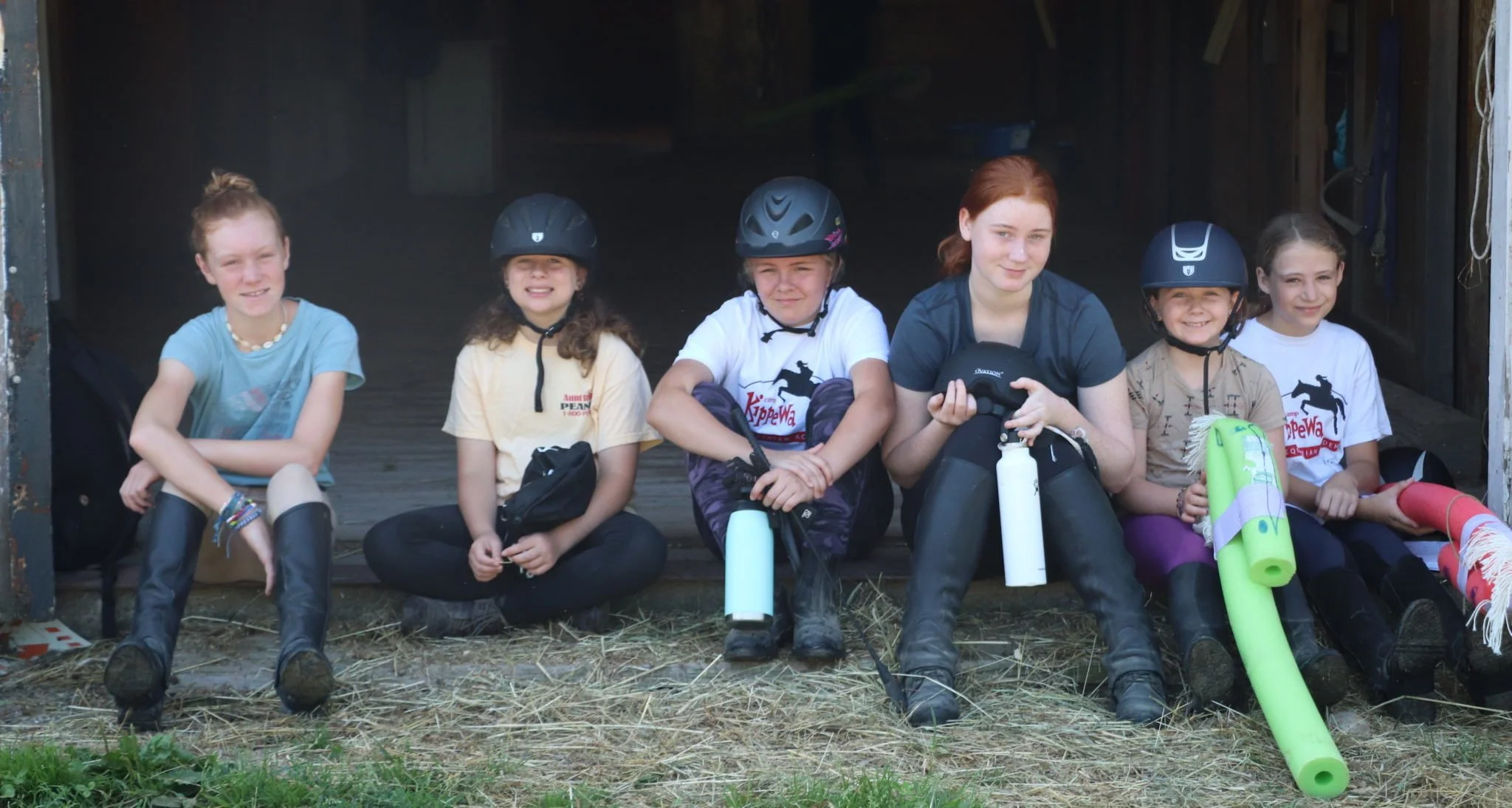 Group of young people sitting in stable wearing riding gear, helmets, and holding pool noodles.